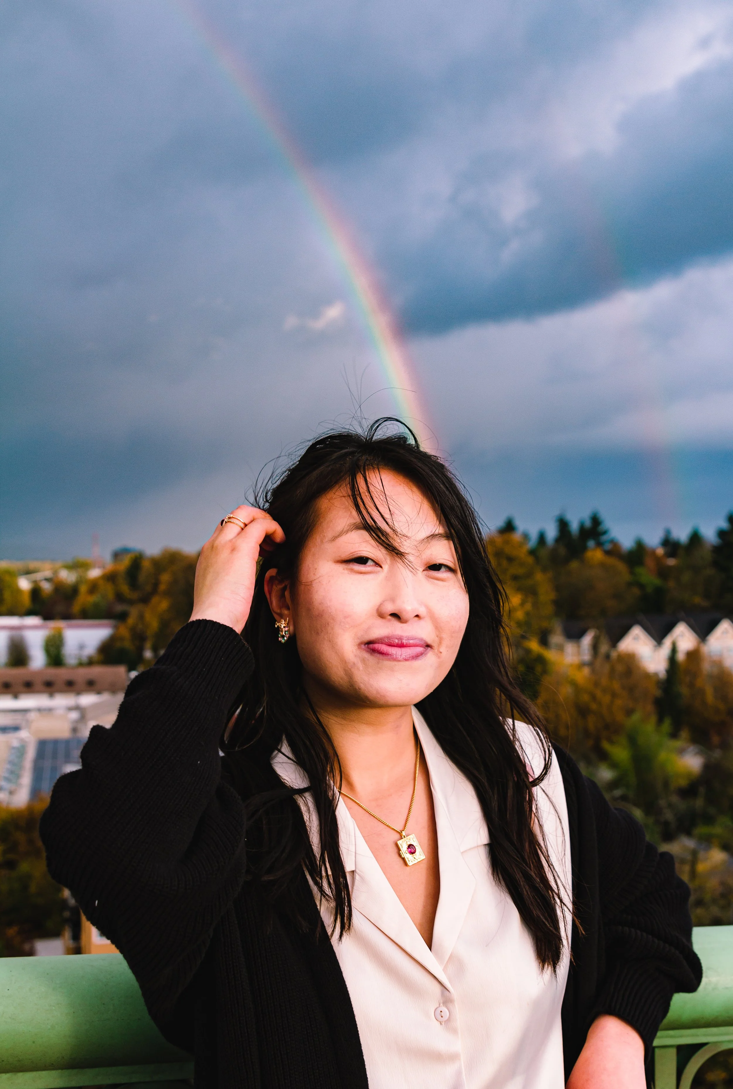 A woman with dark hair and earrings smiling, wearing a beige blouse and black cardigan, standing outdoors with a rainbow in the cloudy sky behind her.
