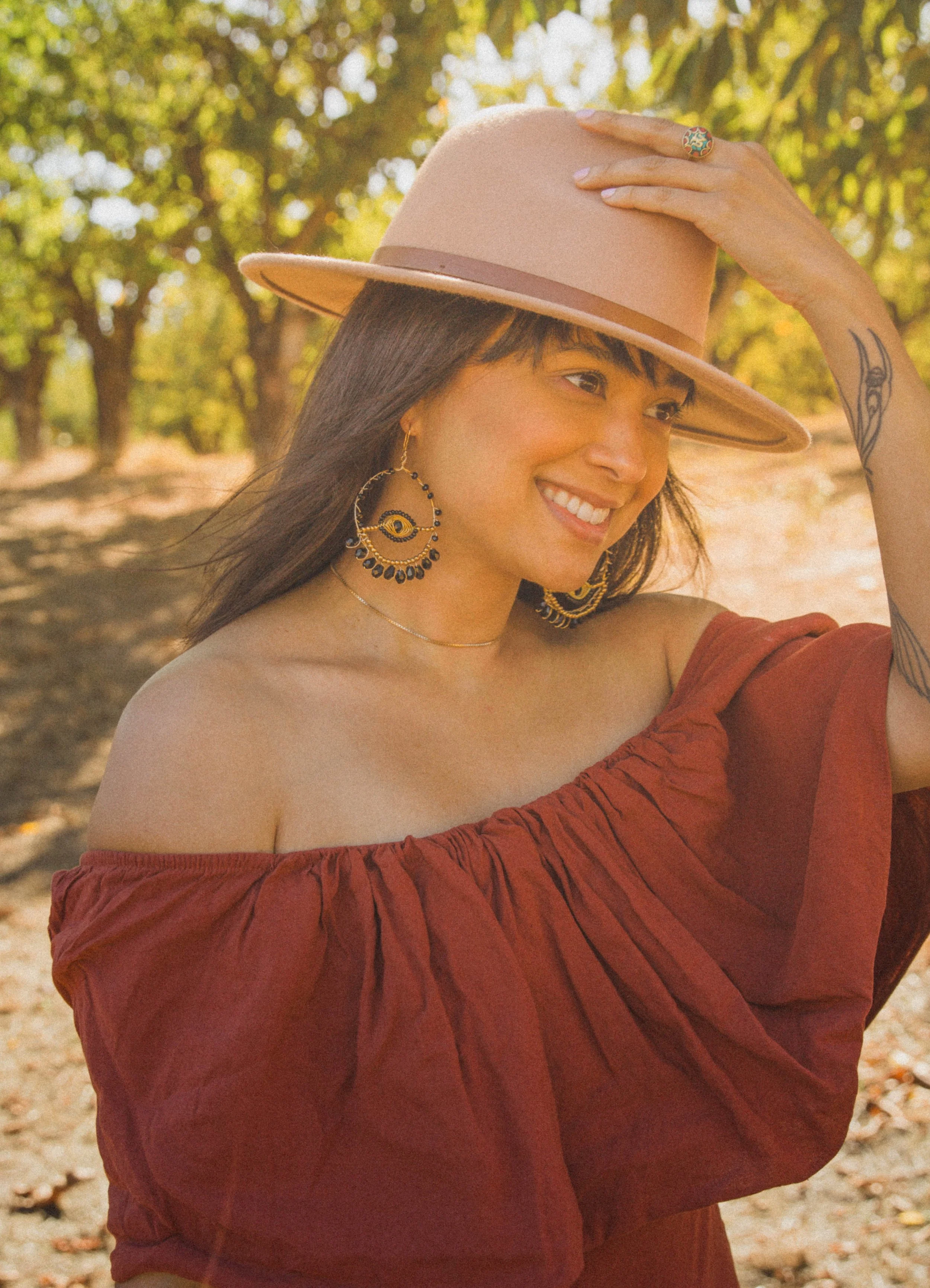 A woman smiling outdoors in a park during fall, wearing a beige hat, red off-shoulder dress, and large jewelry including earrings and a ring, with tattoos on her arm.