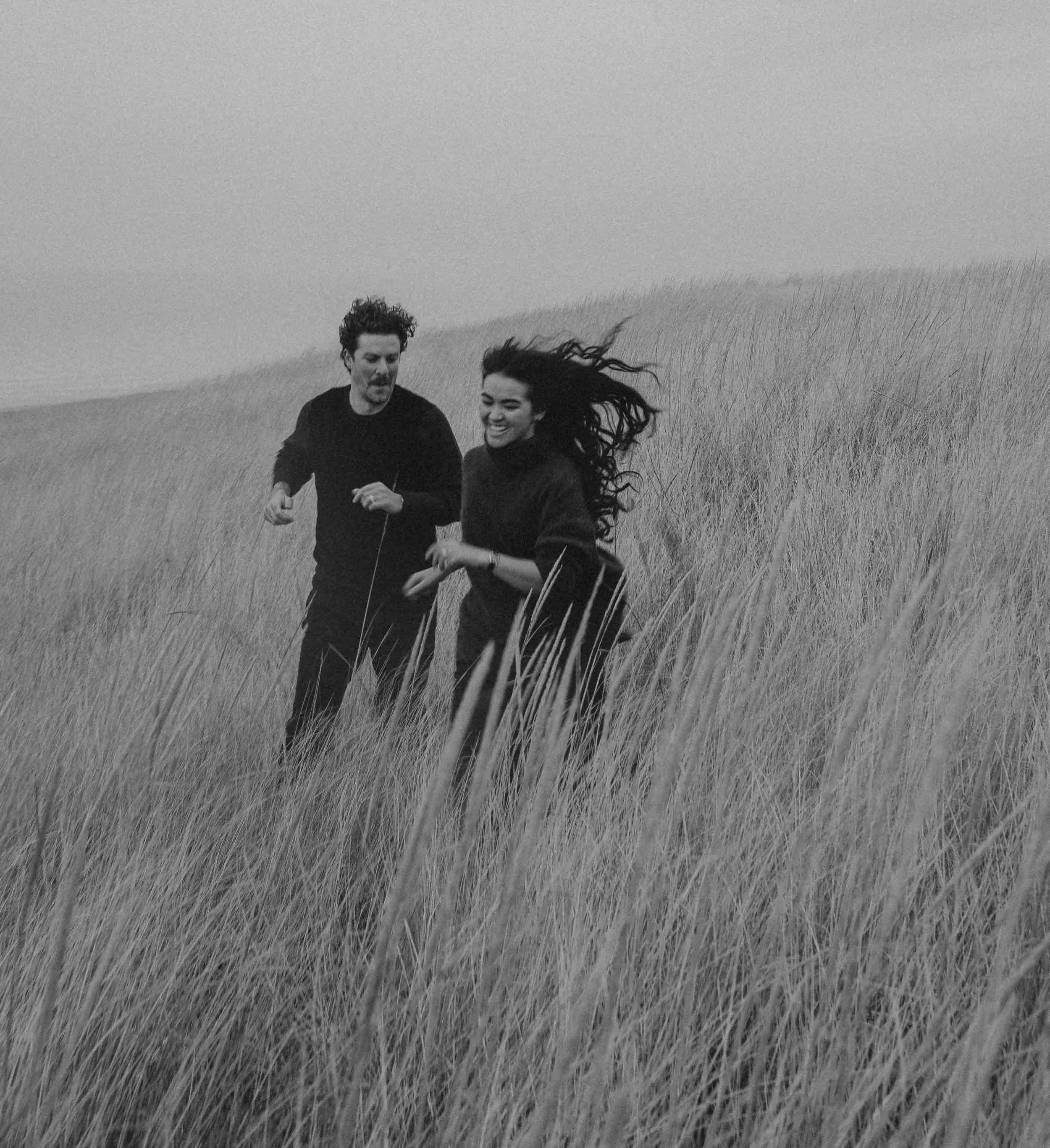 Two people, a man and a woman, running through a grassy field on a cloudy day. The woman has long, wavy hair flowing behind her, and both are smiling.