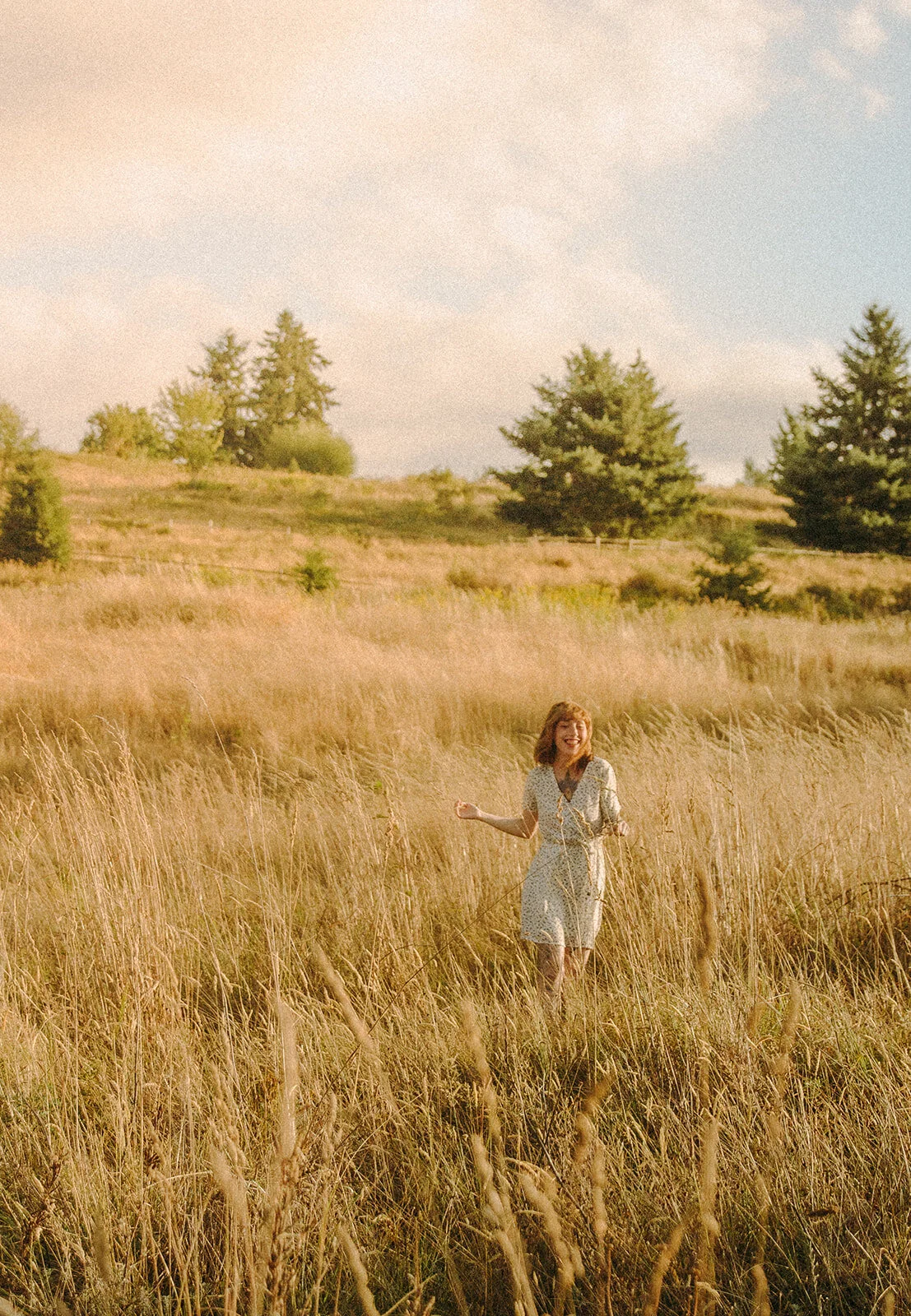A woman in a white dress walking through a field of tall, golden grass with trees in the background and a partly cloudy sky.