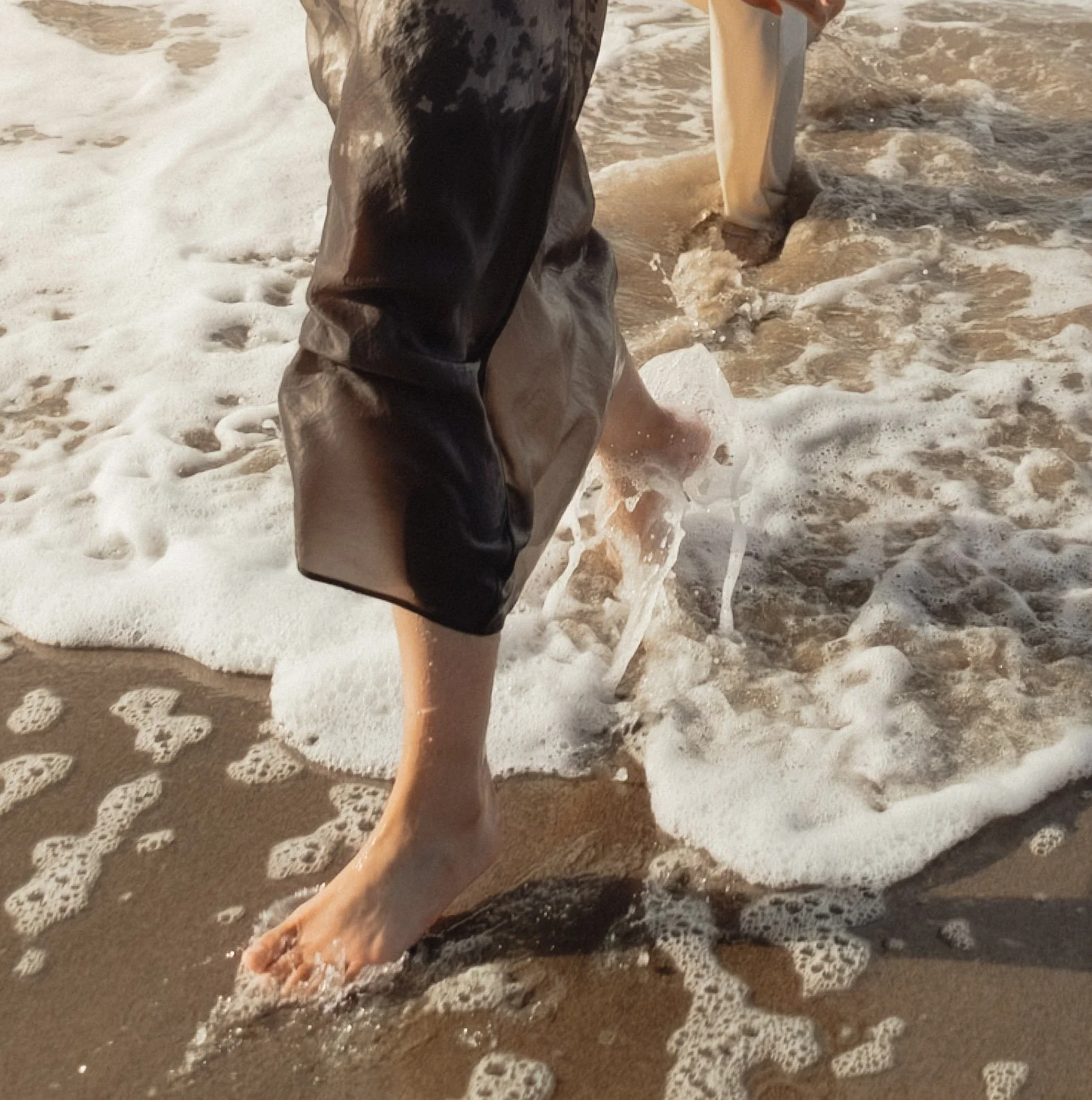 Close-up of a person walking barefoot in the ocean with seashells in the sand, wearing black and beige shorts, with foamy waves around their feet.