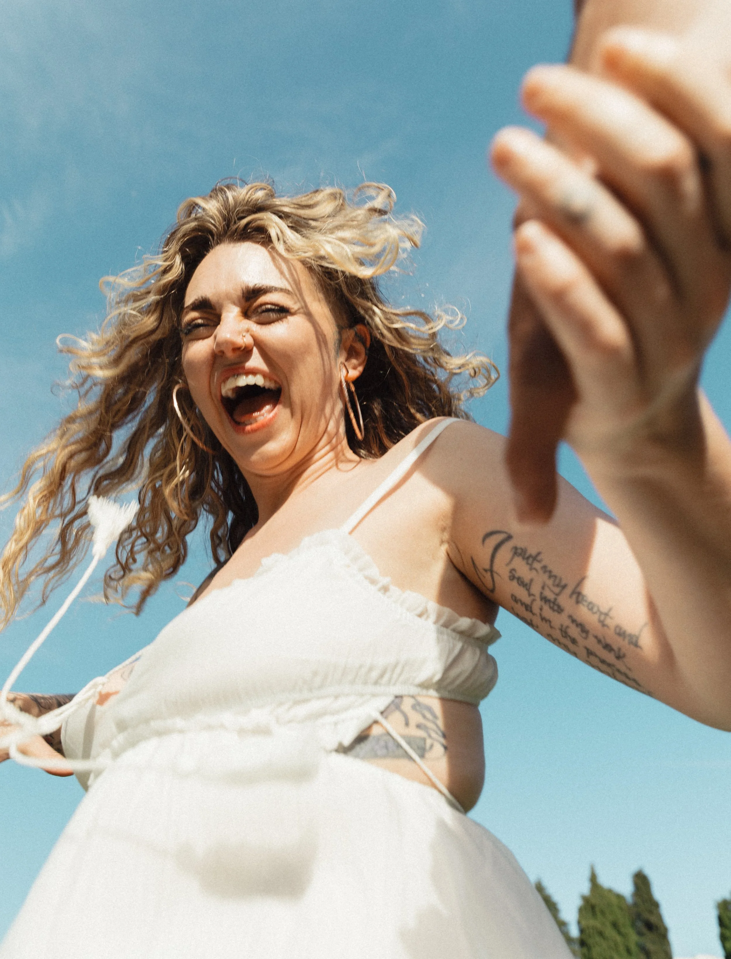 A woman with curly blonde hair wearing a white dress, smiling widely, taking a selfie outside against a clear blue sky.