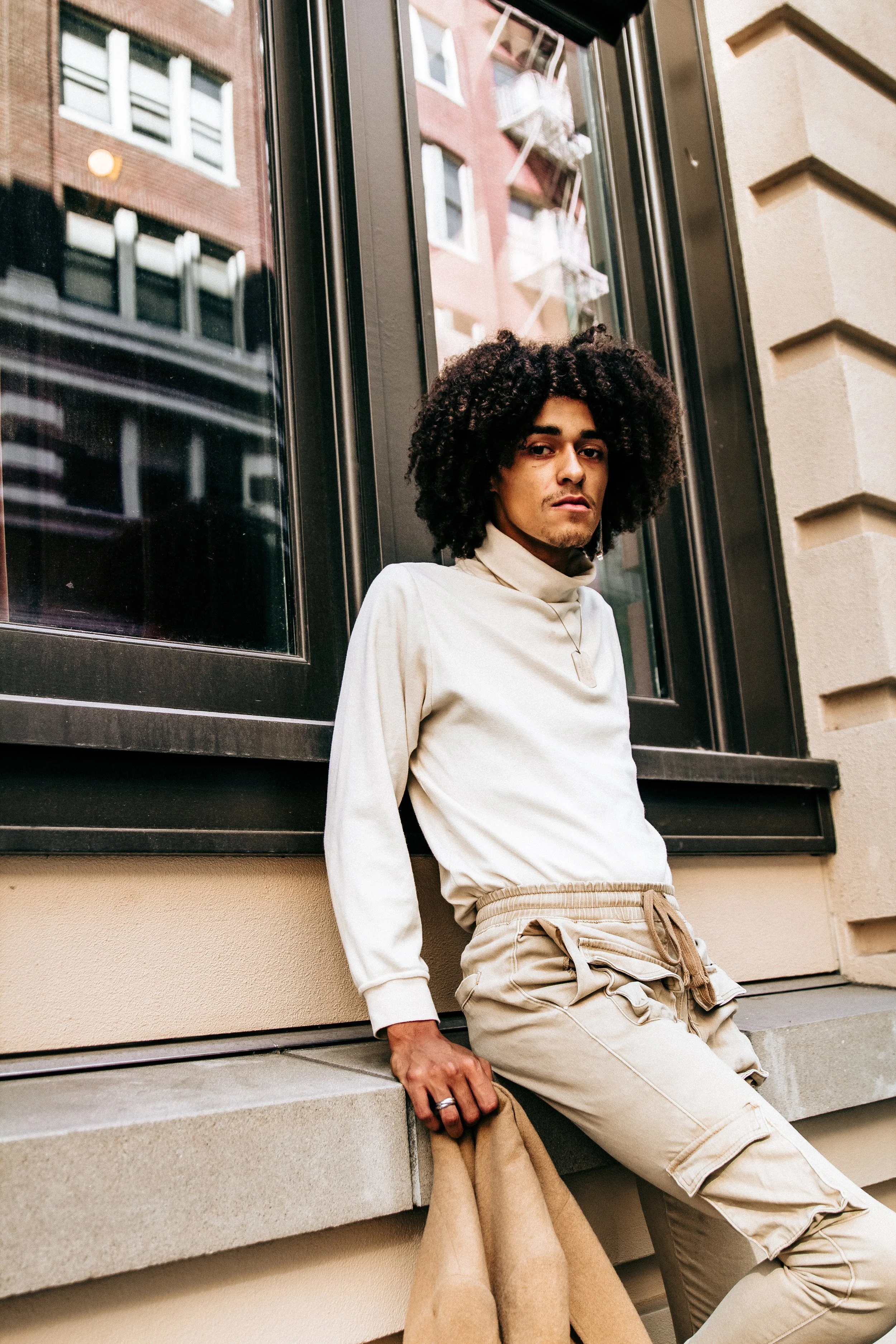 A person with curly hair leaning against a building with windows, wearing a beige sweatshirt and beige cargo pants, with a beige coat hanging from their hand.