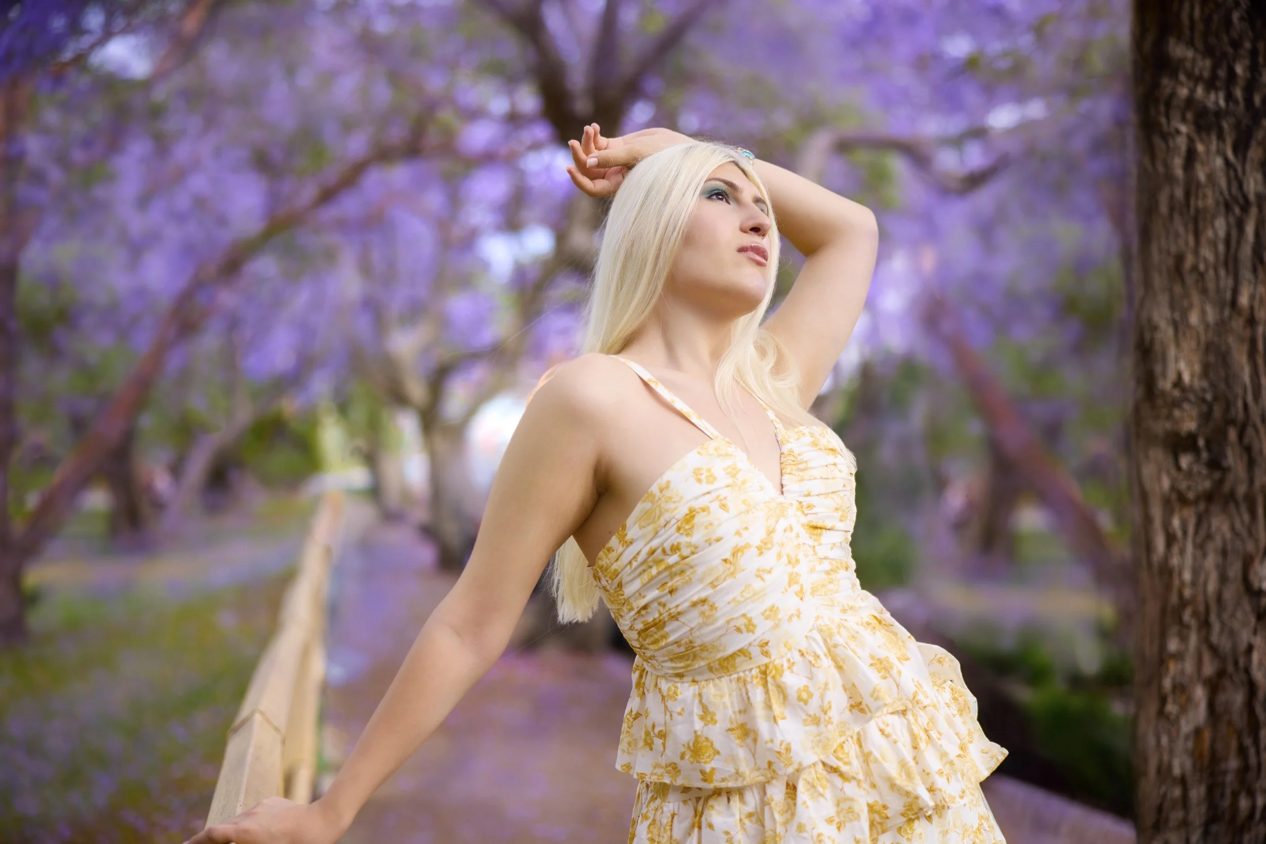 Woman with long blonde hair in a summer dress posing outdoors with purple-blossomed trees in the background.