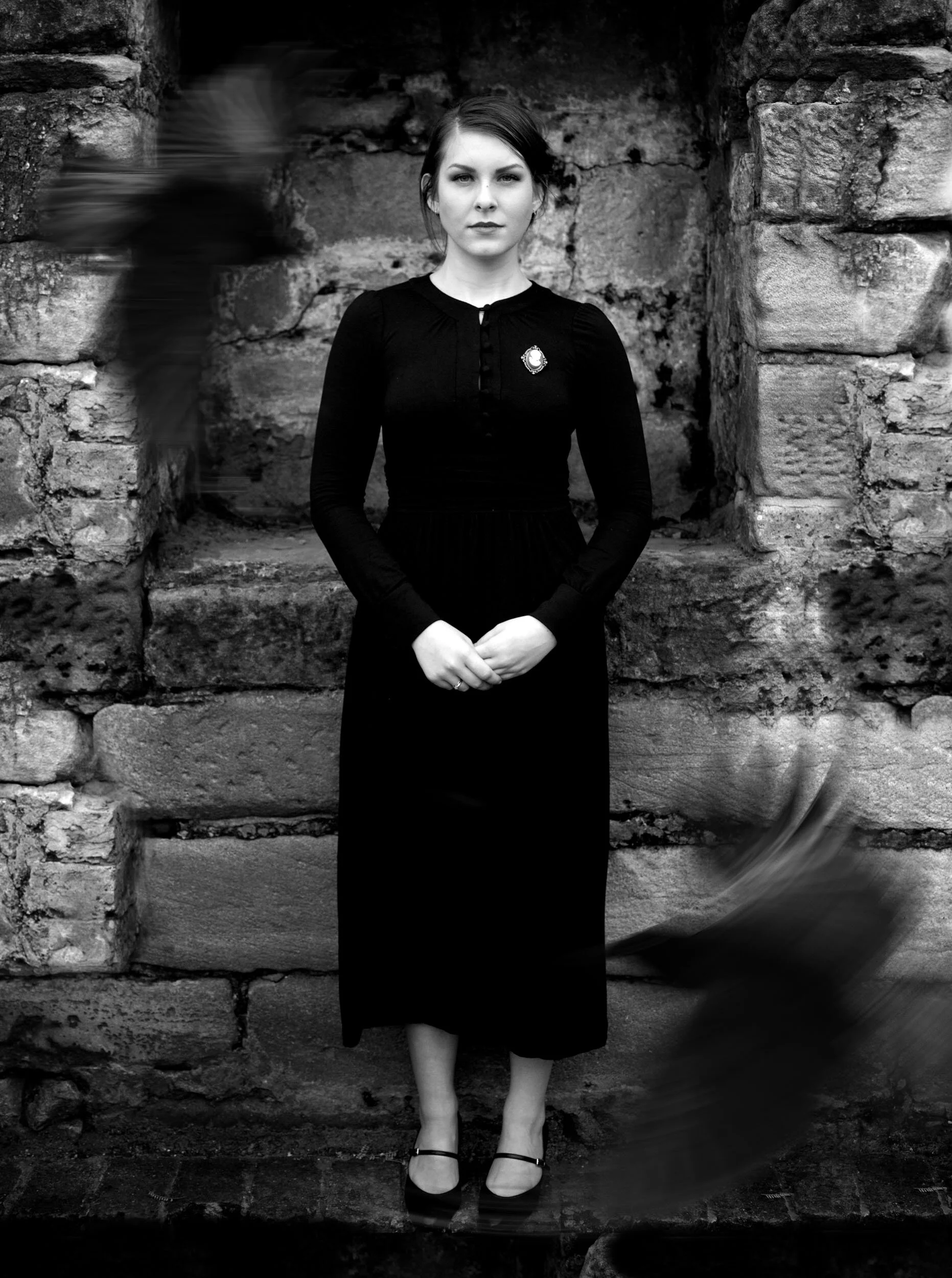 Black and white photo of a woman in a long black dress standing against a stone wall with blurred birds flying around her.