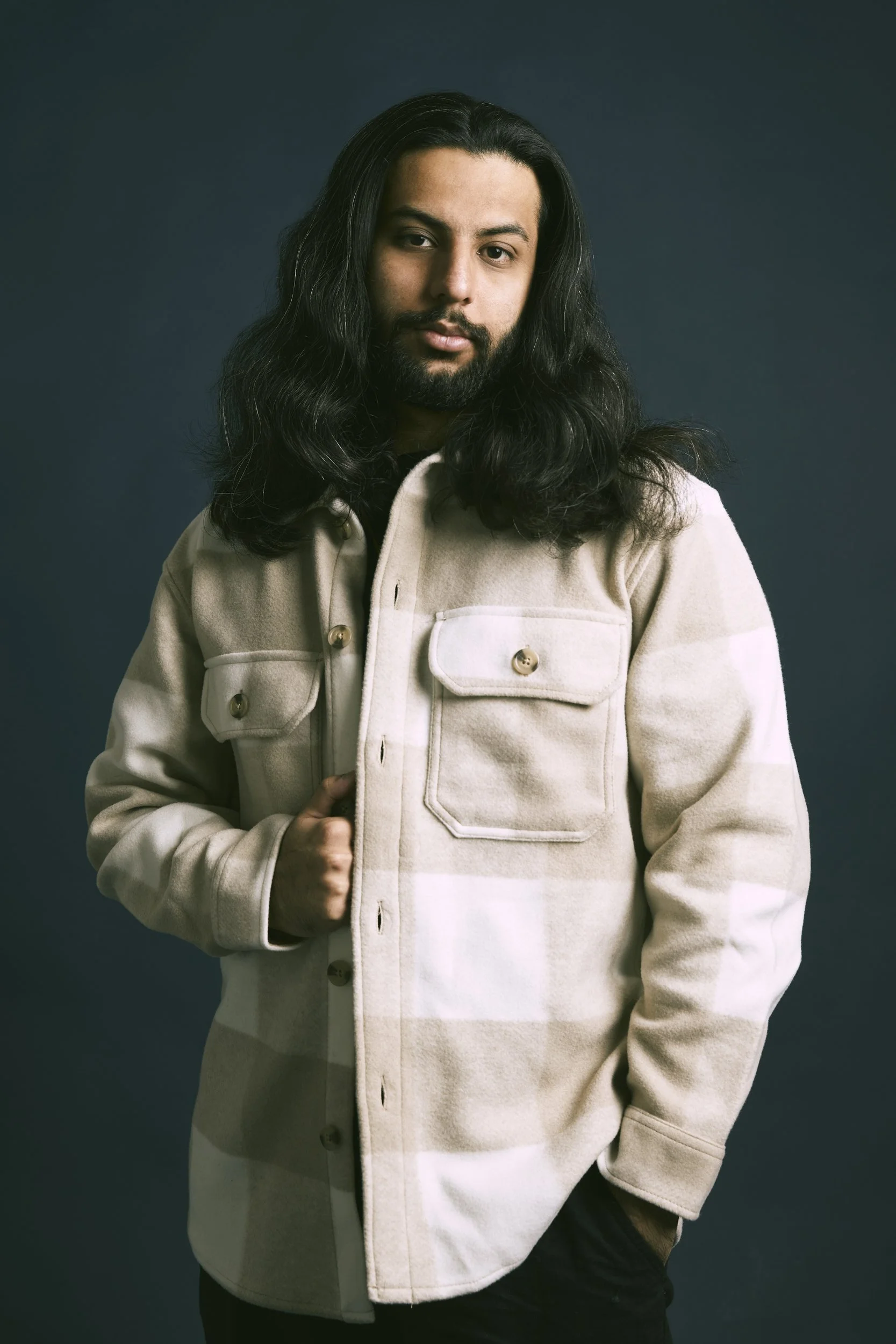 Man with long hair and beard wearing a beige checkered jacket against a dark background.