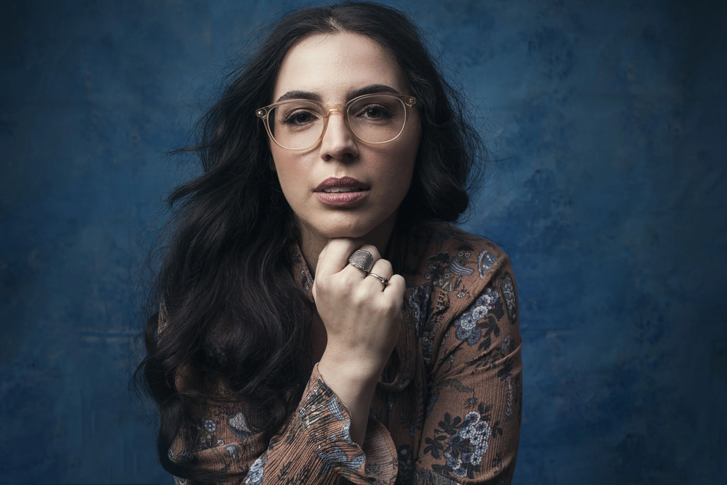 Woman with long dark hair and glasses, wearing a patterned blouse, against a blue background.