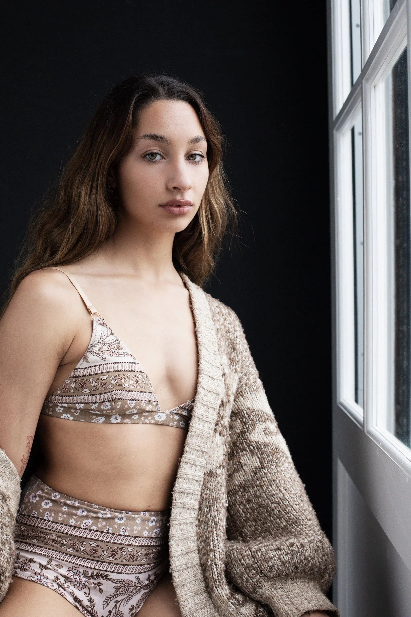 Woman wearing a patterned bikini and a knitted cardigan, standing by a window with a dark background.