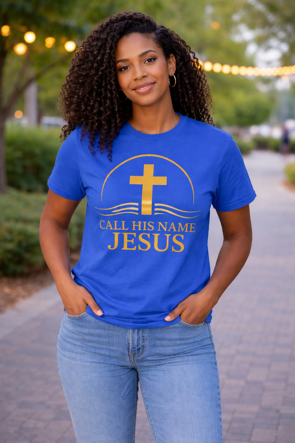 Woman wearing blue “Call His Name Jesus” t-shirt by Michelle Shearer with gold design in outdoor setting
