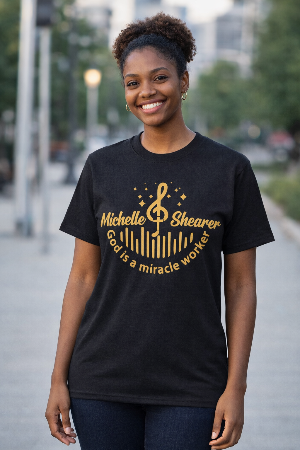 Woman wearing black “God is a miracle worker” t-shirt by Michelle Shearer with gold design in outdoor setting