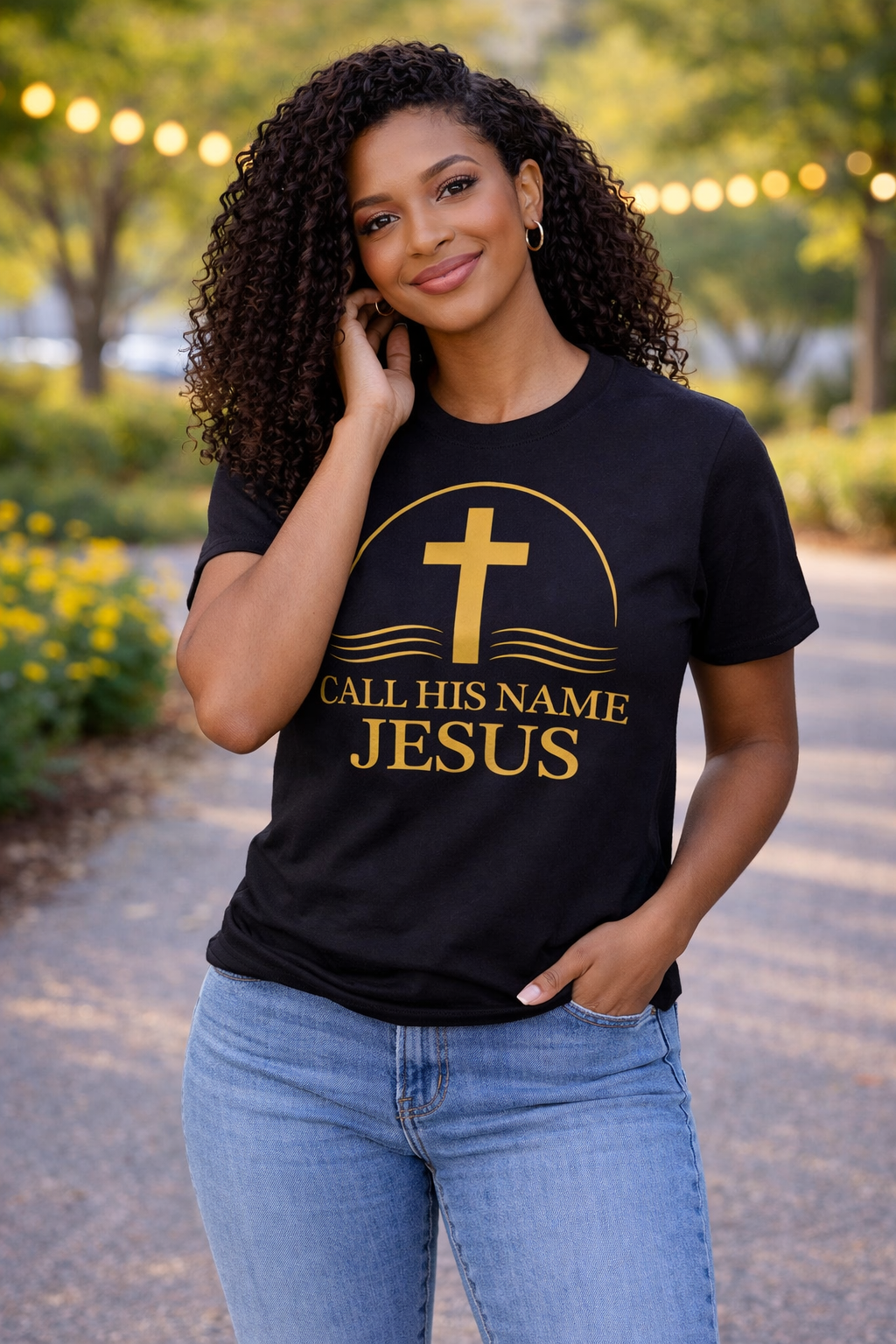 Woman wearing black “Call His Name Jesus” t-shirt by Michelle Shearer with gold design in outdoor park setting