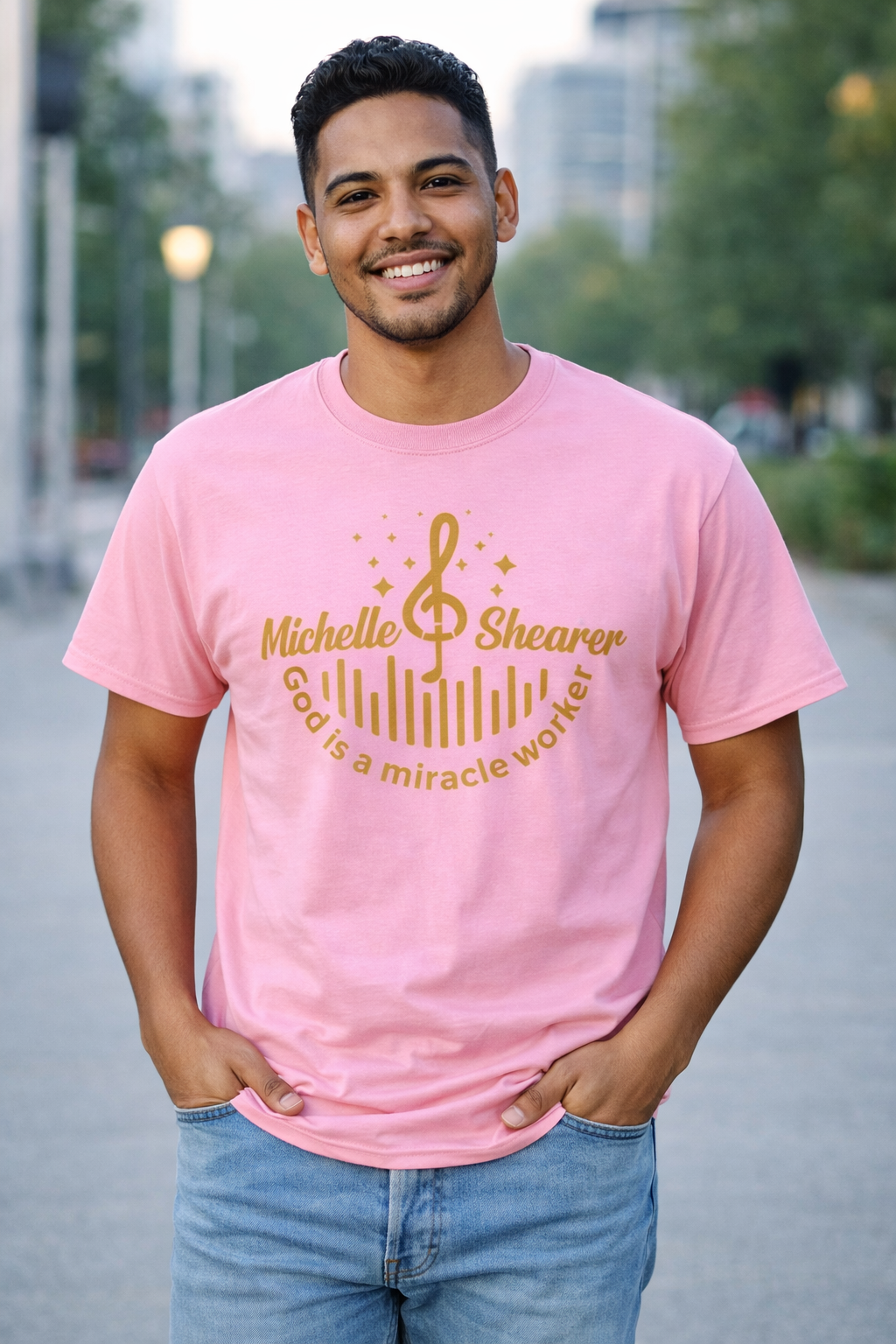 Man wearing pink “God is a miracle worker” t-shirt by Michelle Shearer with gold design in outdoor setting