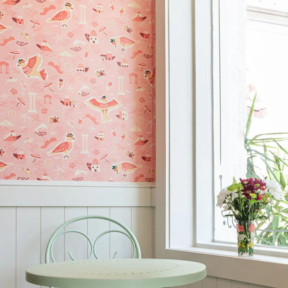 A pastel-colored interior with a pink wallpaper featuring whimsical illustrated birds, animals, and cottages, a white round table with a pastel green chair, and a window with a vase of fresh flowers on the windowsill.