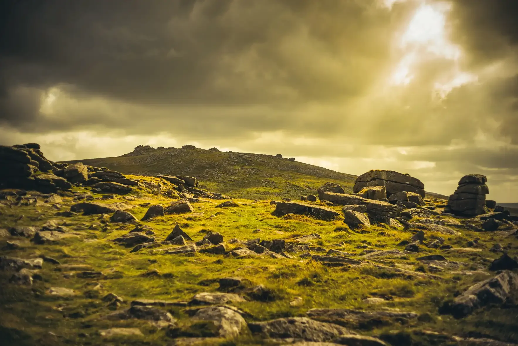 A rocky landscape with a grassy terrain and large stones and boulders. A hill or mountain is visible in the background under a cloudy sky with sunlight breaking through.