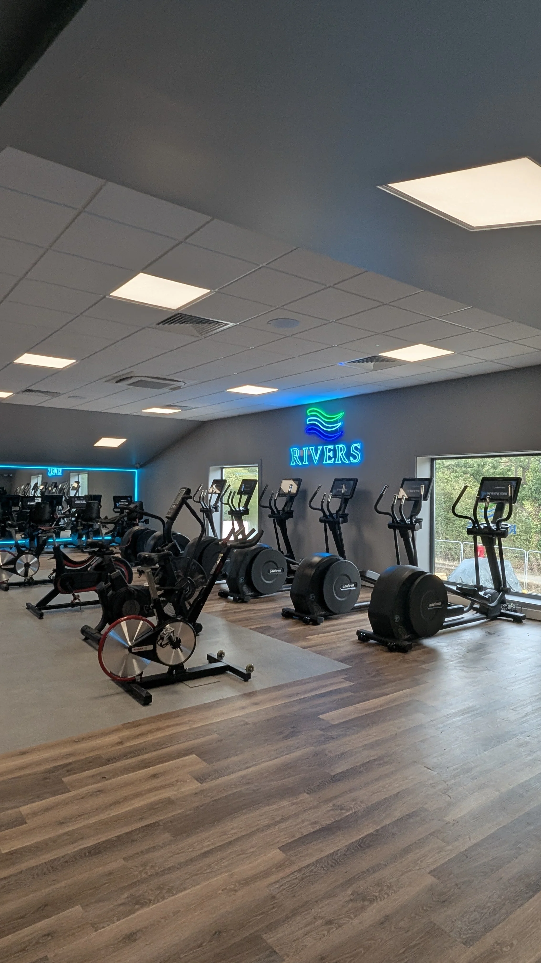 Row of treadmills with digital screens in a gym, grey wall with neon Rivers sign above. Large mirror with blue LED lights around.