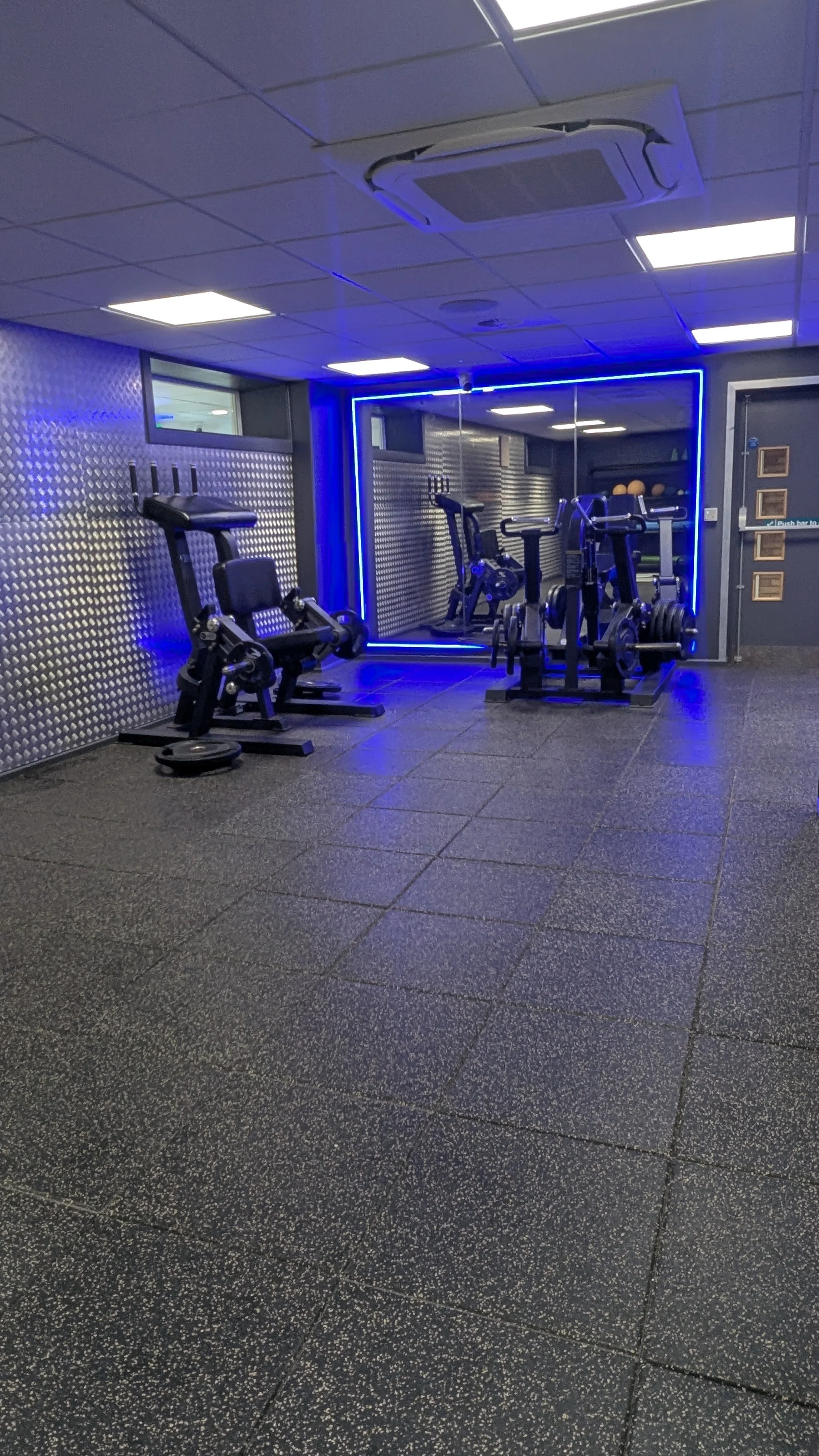 Indoor gym area with a large mirror with blue LED lights, soft black floor, grey and silver metal walls.