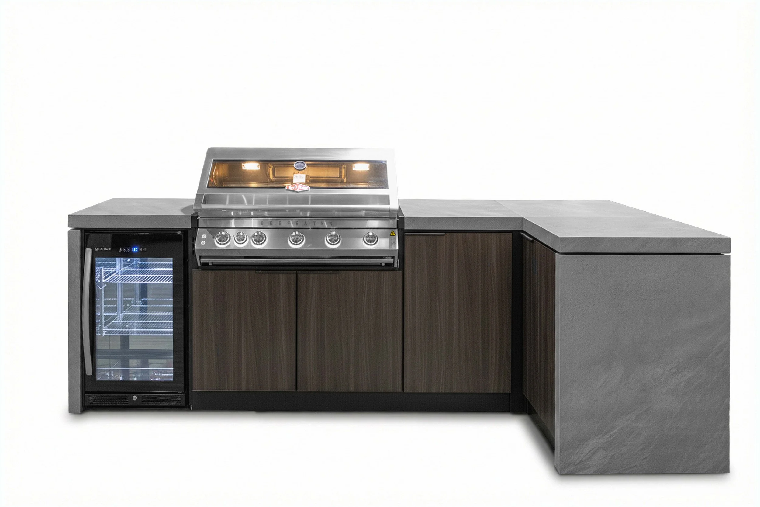 A commercial-grade stainless steel barbeque grill with five control knobs and a small warming rack, positioned on a wooden cabinet with a countertop, next to a beverage refrigerator, against a plain white wall.