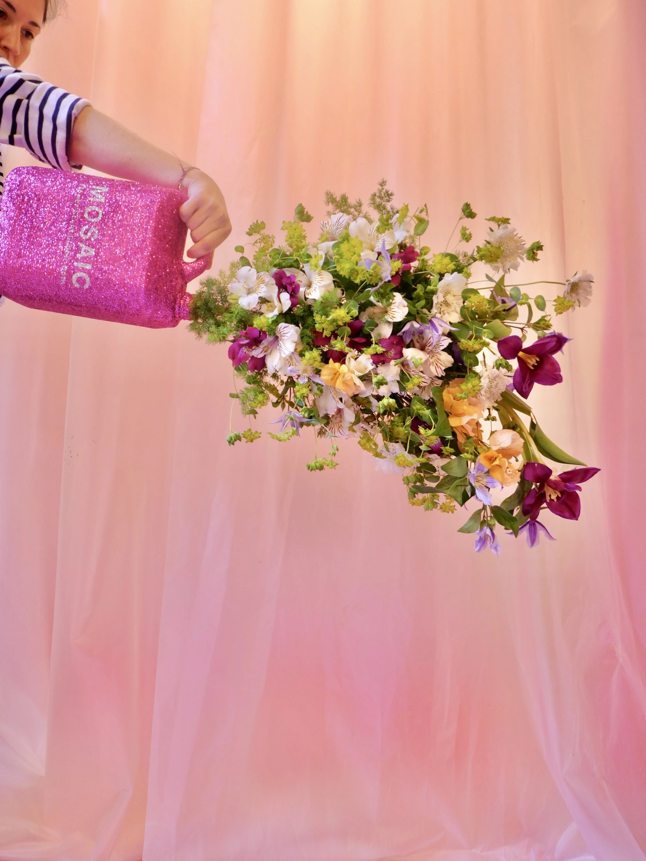 Hand holding Mosaic’s pink bottle with colorful flowers pouring out, designed for The Bathroom immersive exhibition
