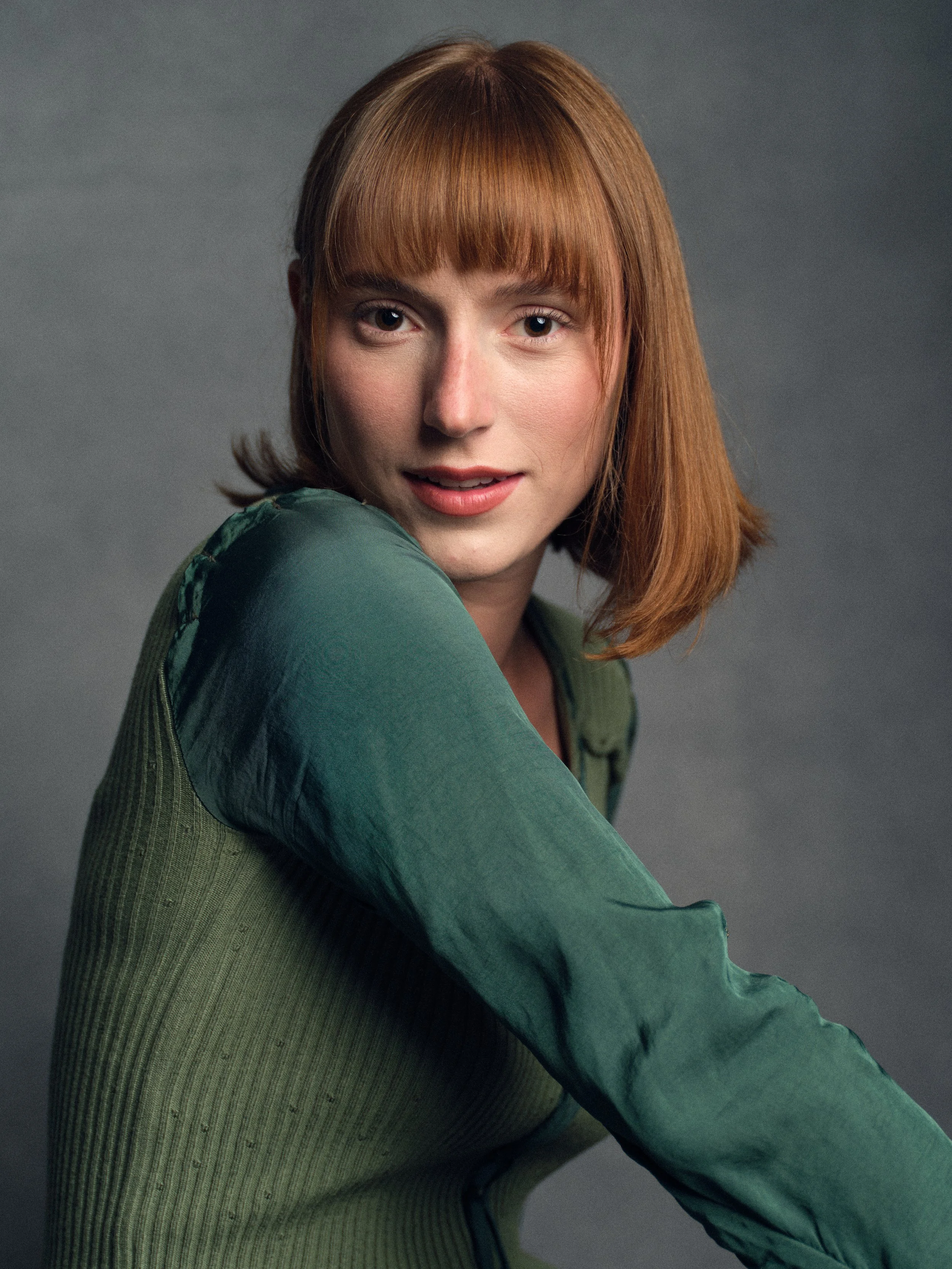 A woman with shoulder-length red hair and bangs, wearing a green textured jacket, looking at the camera with a slight smile against a gray background.