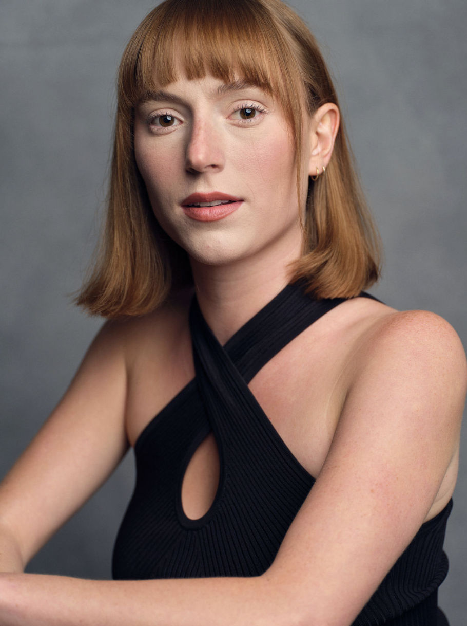 Portrait of a young woman with red hair and fair skin wearing a black sleeveless top, looking directly at the camera against a gray background.