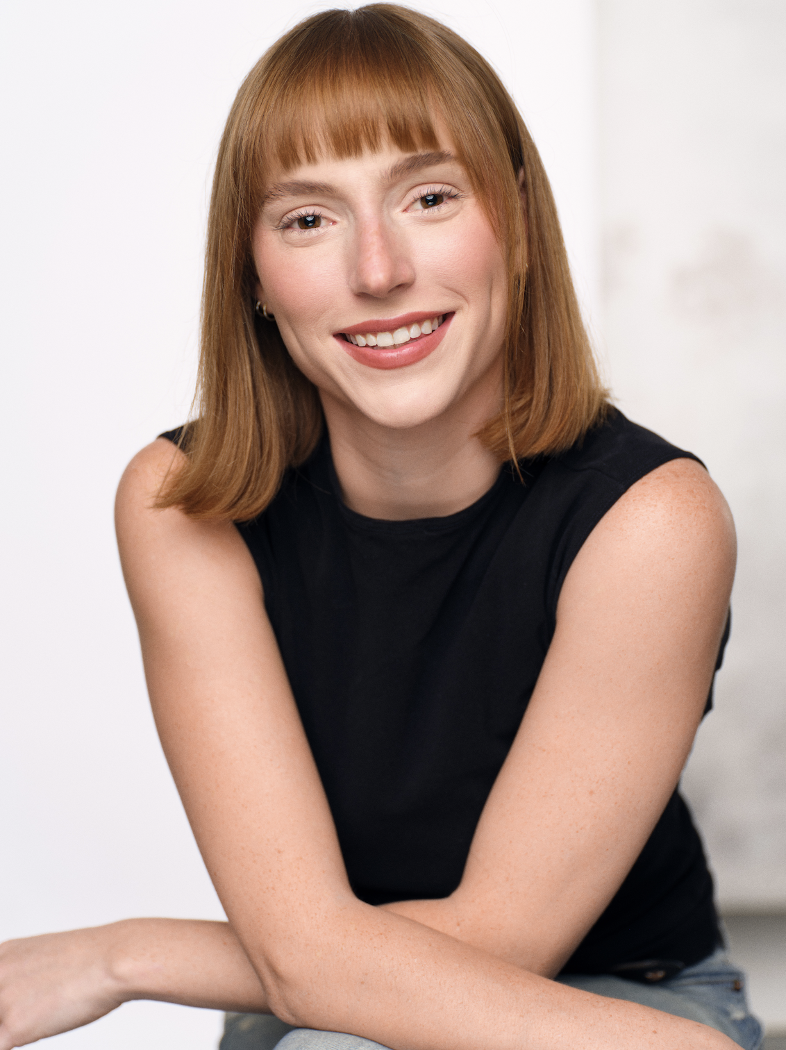 A young woman with shoulder-length red hair, wearing a black sleeveless top, smiling and sitting with her arms crossed.