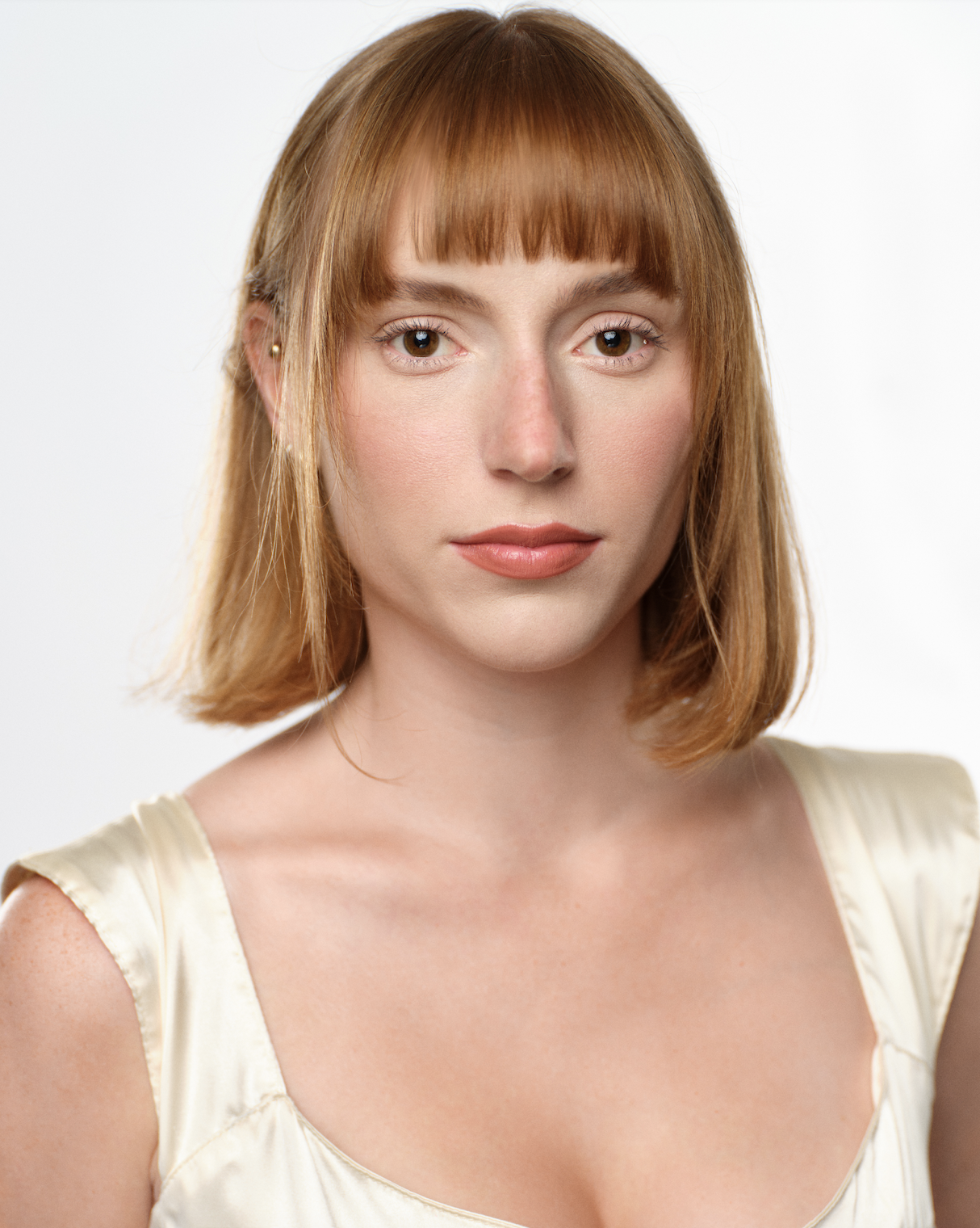 Close-up portrait of a young woman with shoulder-length auburn hair and bangs, wearing a cream-colored satin top against a plain white background.