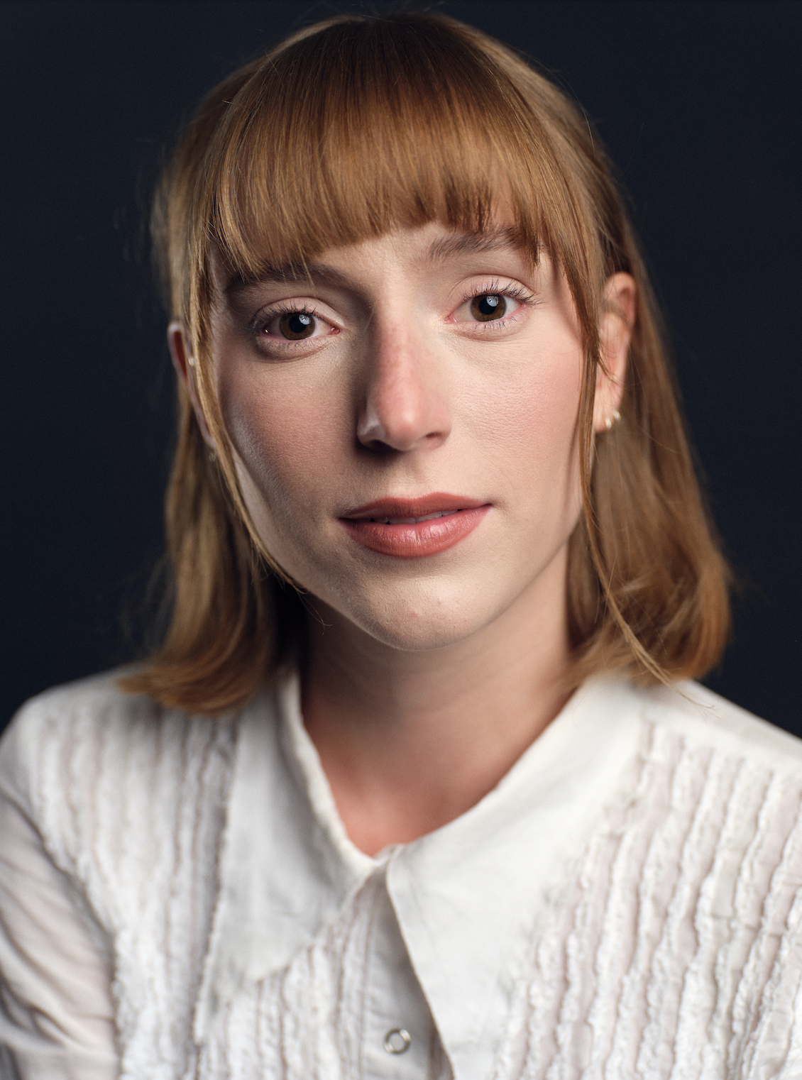 A woman with red hair and light makeup, wearing a white textured blouse, against a dark background.