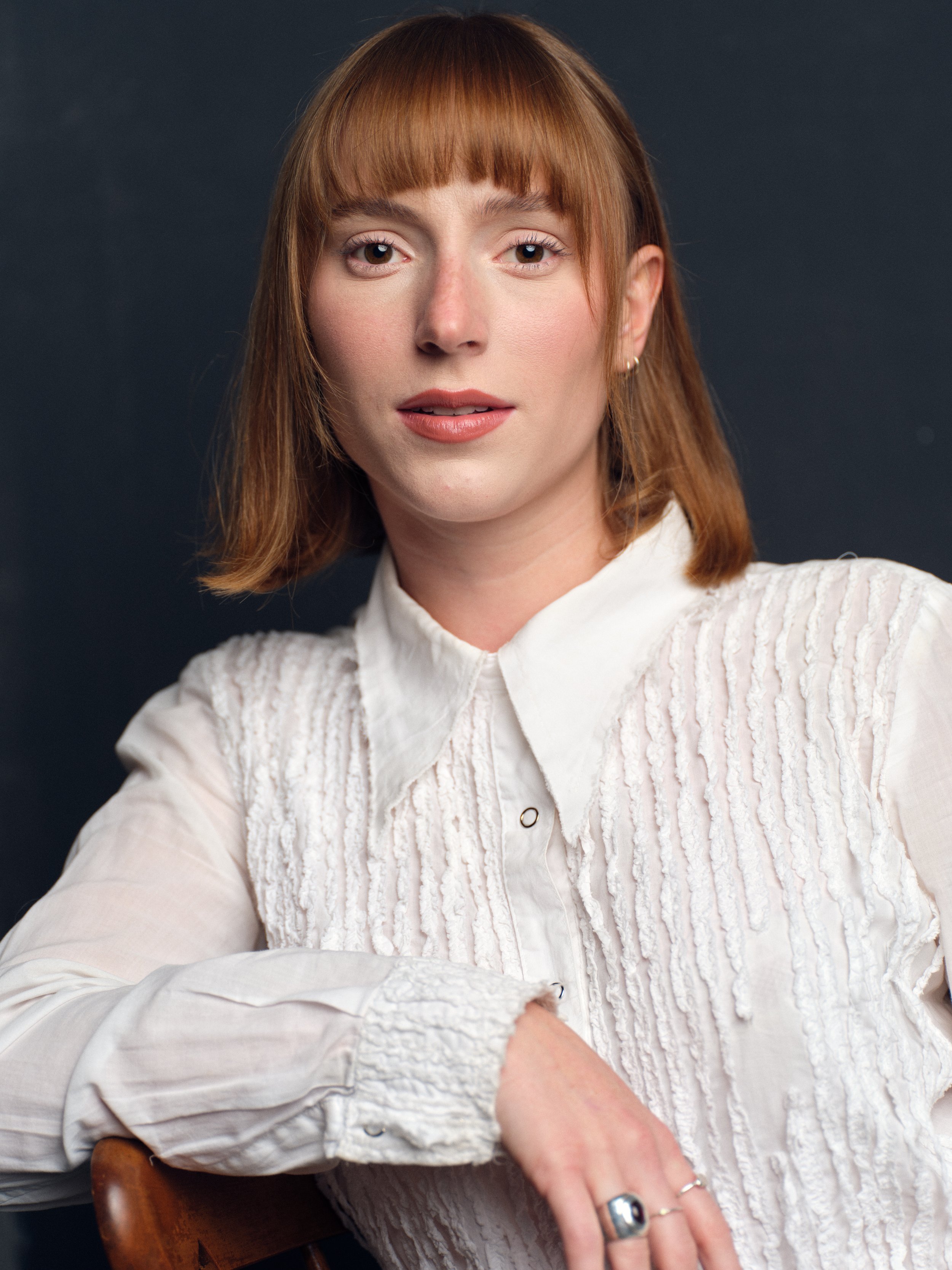 A woman with light skin and reddish-brown hair in a bob haircut, wearing a white textured blouse with a high collar, sitting with her arm resting on a wooden chair, against a dark background.