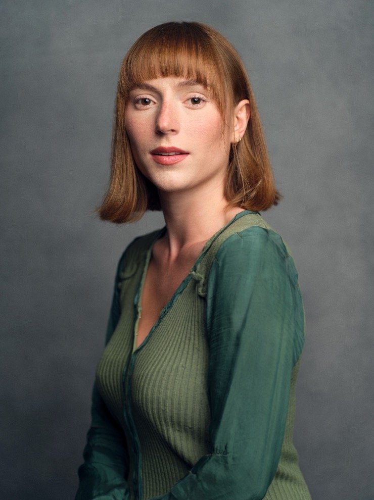 Portrait of a woman with shoulder-length reddish hair and bangs, wearing a greenish top, against a gray background.