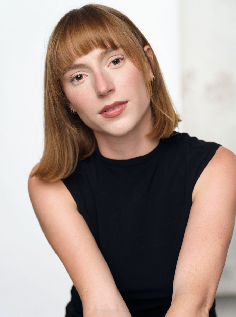 Portrait of a young woman with shoulder-length red hair, wearing a sleeveless black top, and earrings, with a neutral expression, against a plain light background.