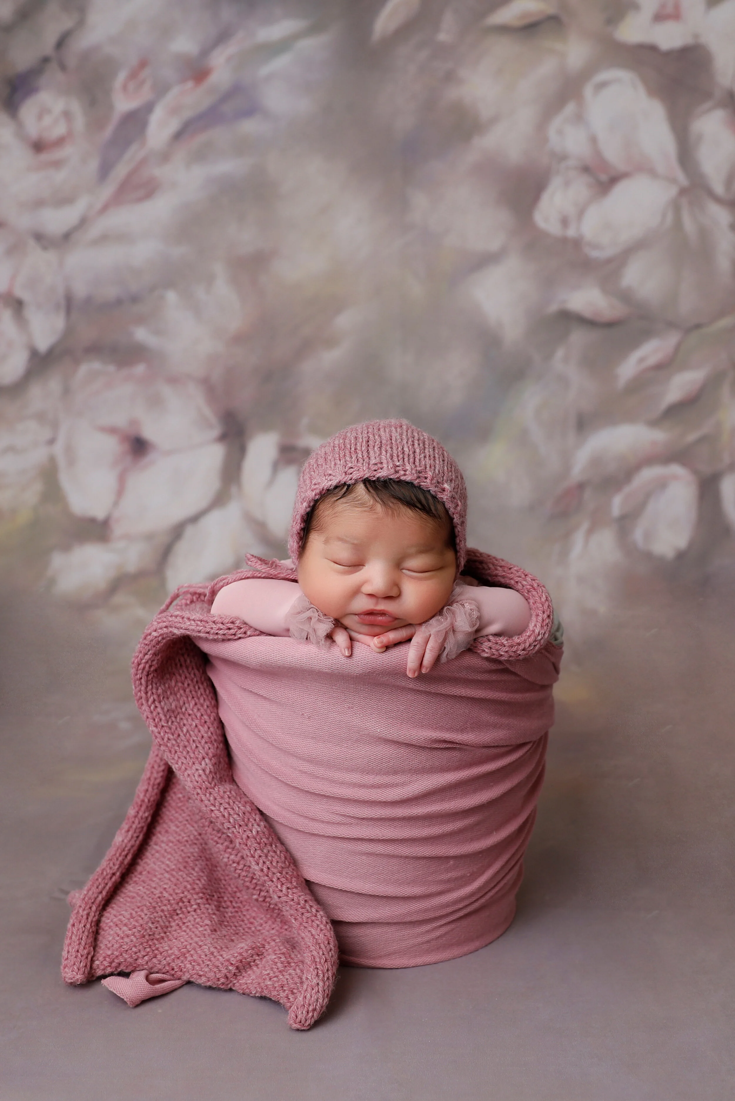 A sleeping newborn baby wrapped in pink fabric, wearing a pink knit hat, against a soft floral background.