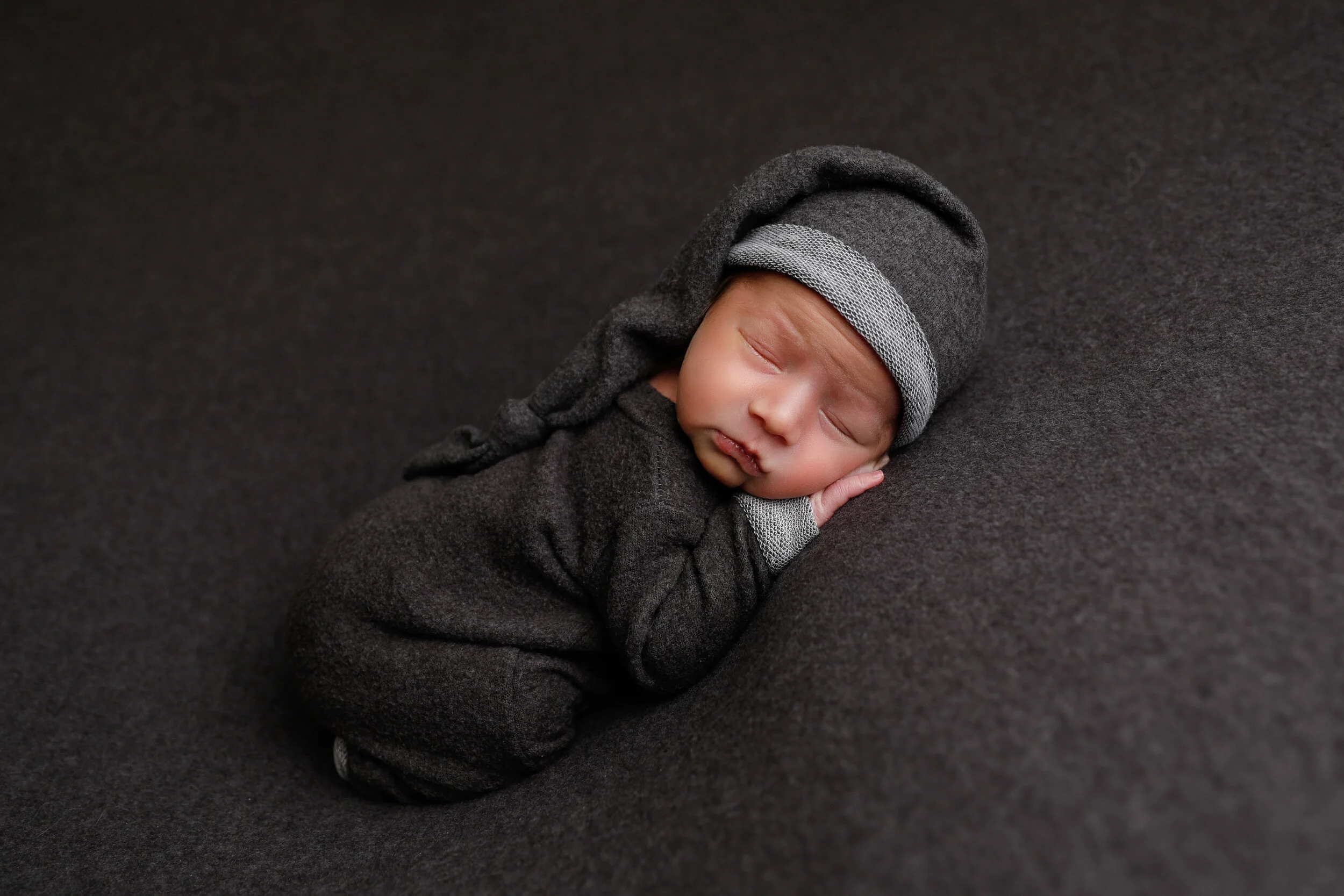 A newborn baby sleeping, dressed in a dark gray outfit and hat, lying on a dark gray surface.
