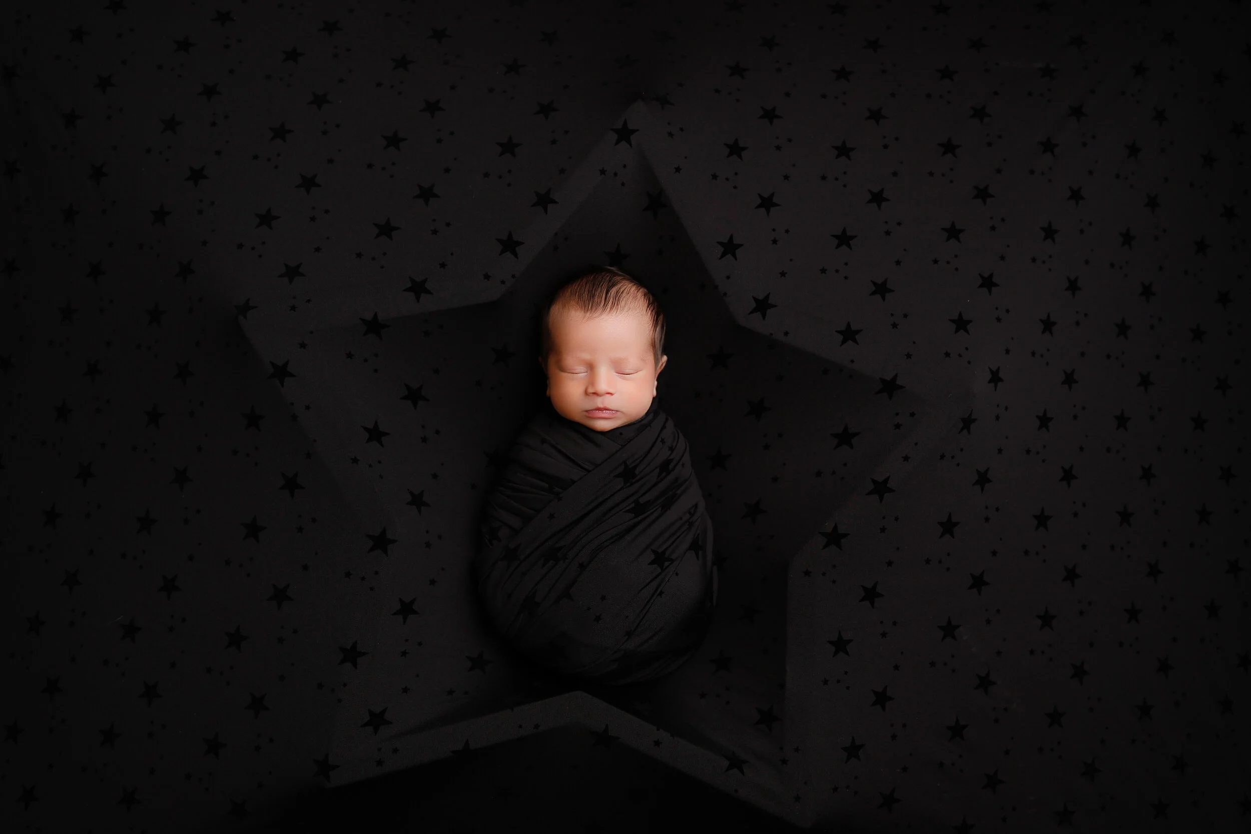 A newborn baby wrapped in a black blanket with star patterns, sleeping inside a 3D star-shaped cutout on a black star-patterned background.