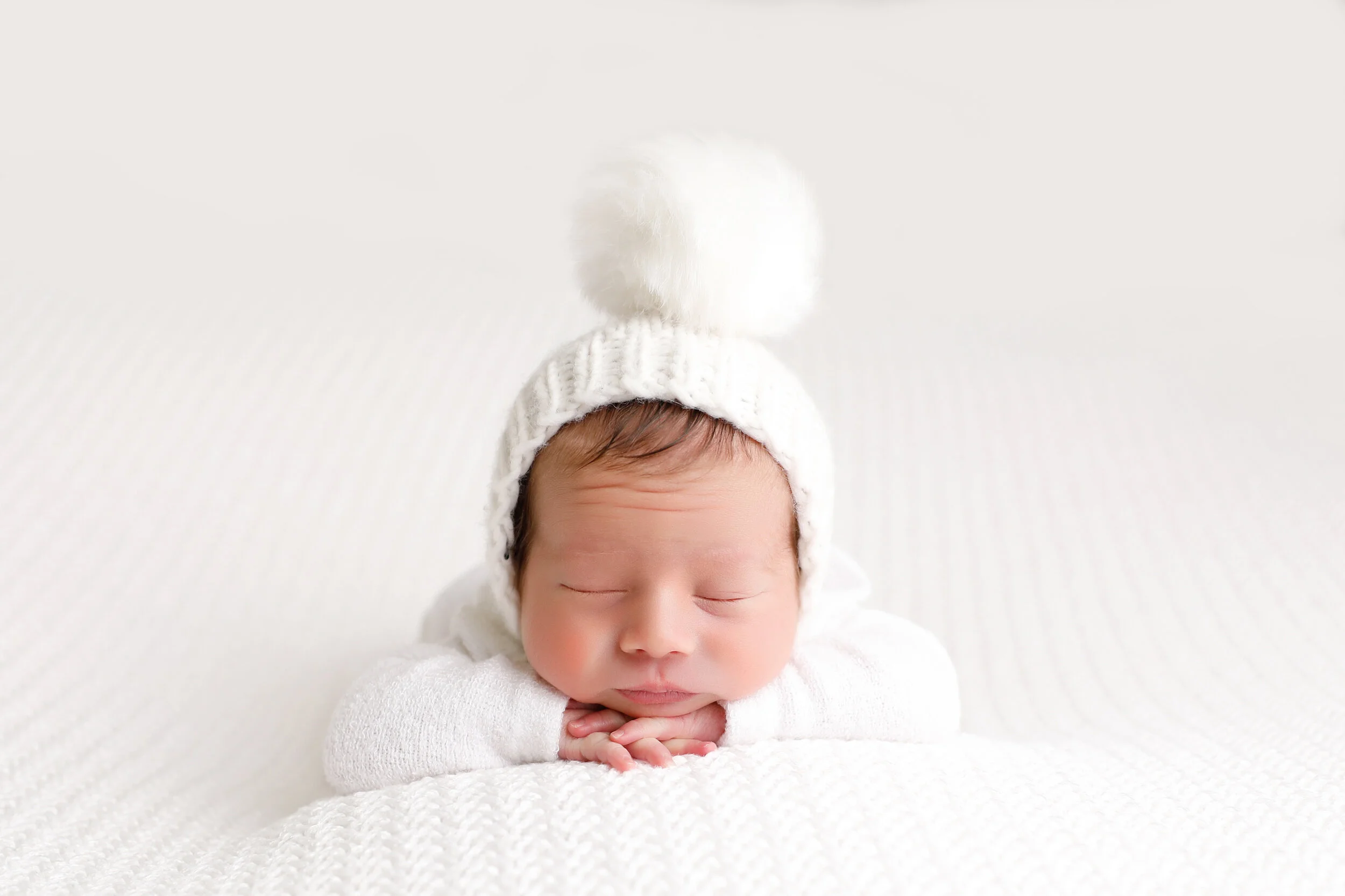 A sleeping baby wearing a white knit hat with a pom-pom, lying on a white textured blanket.