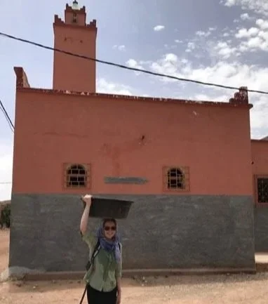 Beth standing in front of a mud-brick building holding a large black bucket on her head.