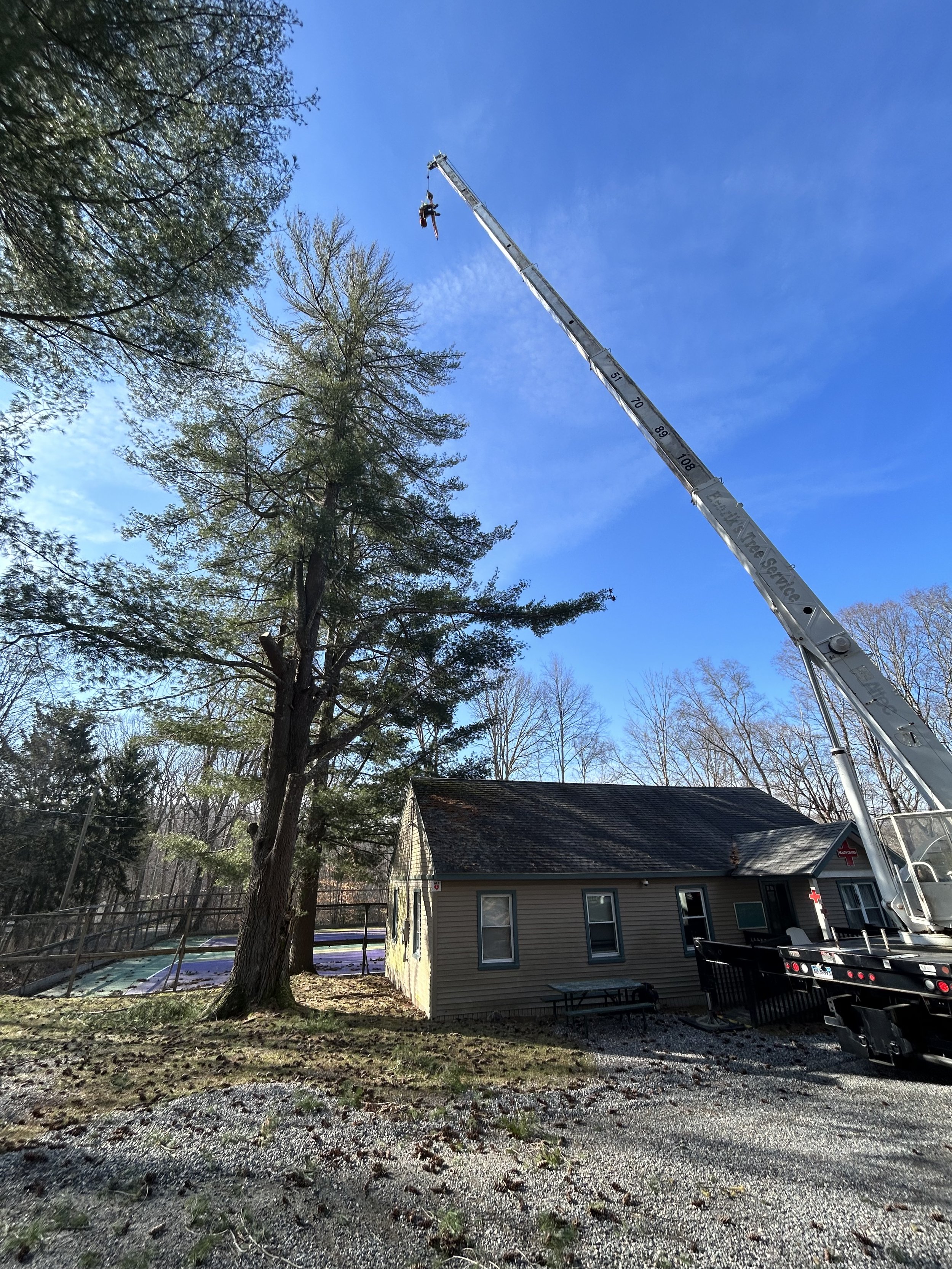 A large crane lifting a worker high into the sky near a residential house with trees in the background.
