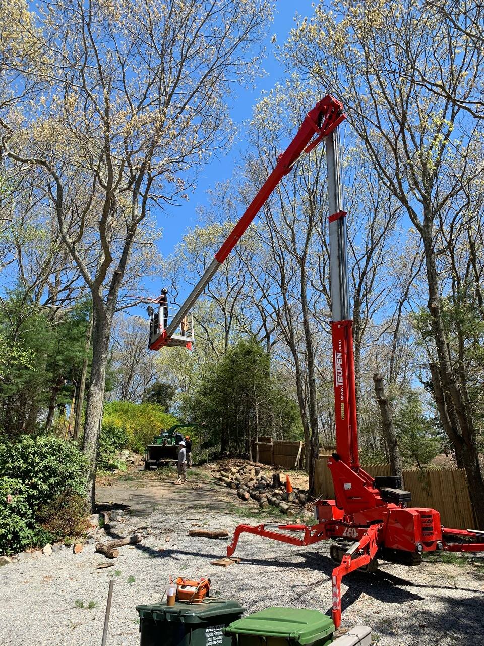 A red telescopic boom lift elevated in a backyard, with two workers operating it among tall trees with budding spring leaves.