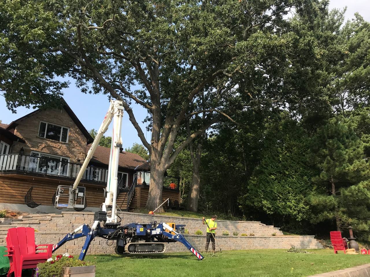 A worker in a yellow vest operating a tree trimming robot with a large arm, near a large tree in a landscaped backyard with brick steps and red chairs.