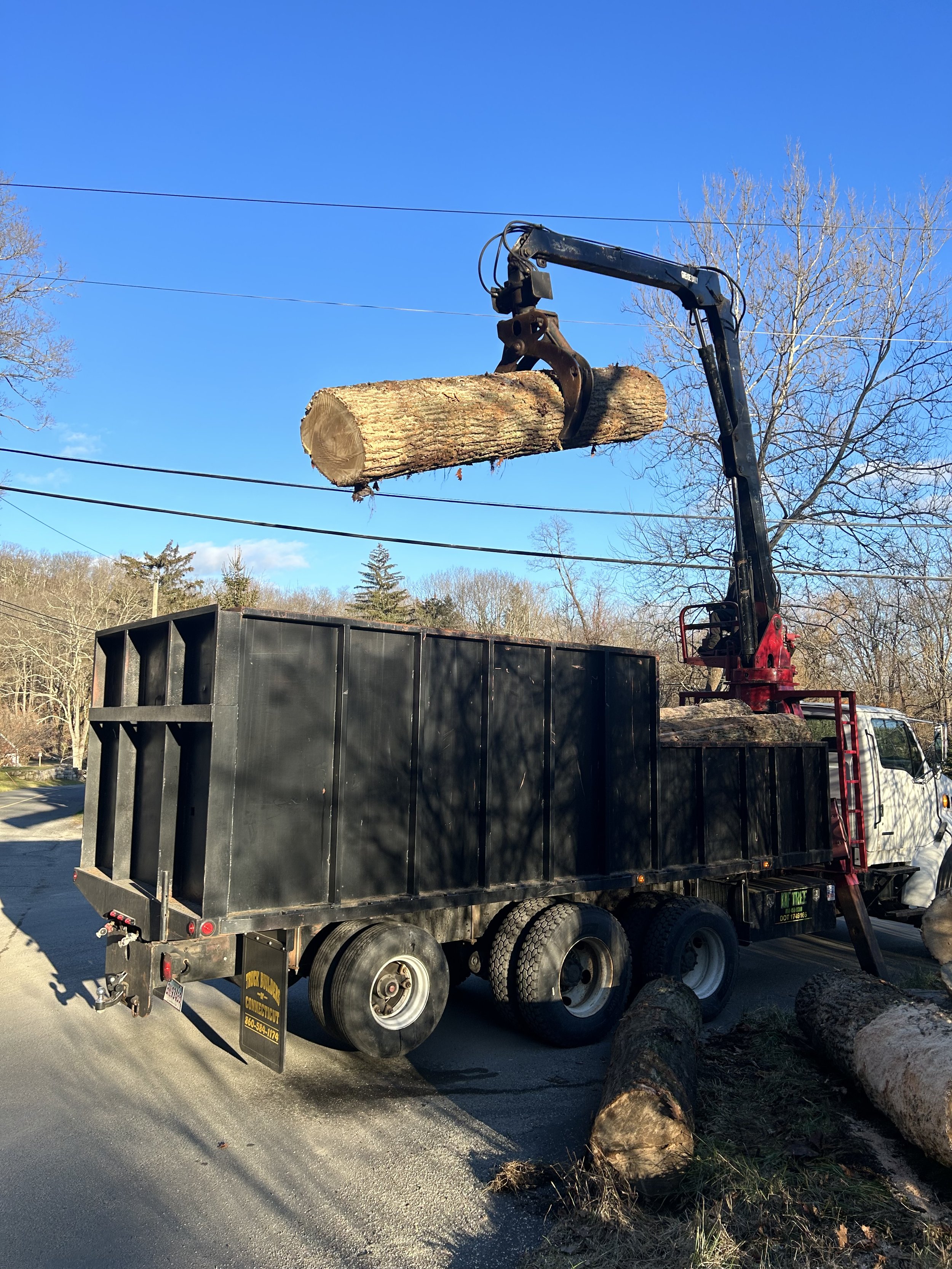 A logging truck with a mechanical arm lifting a large log, parked on a street with fallen logs nearby and power lines overhead.