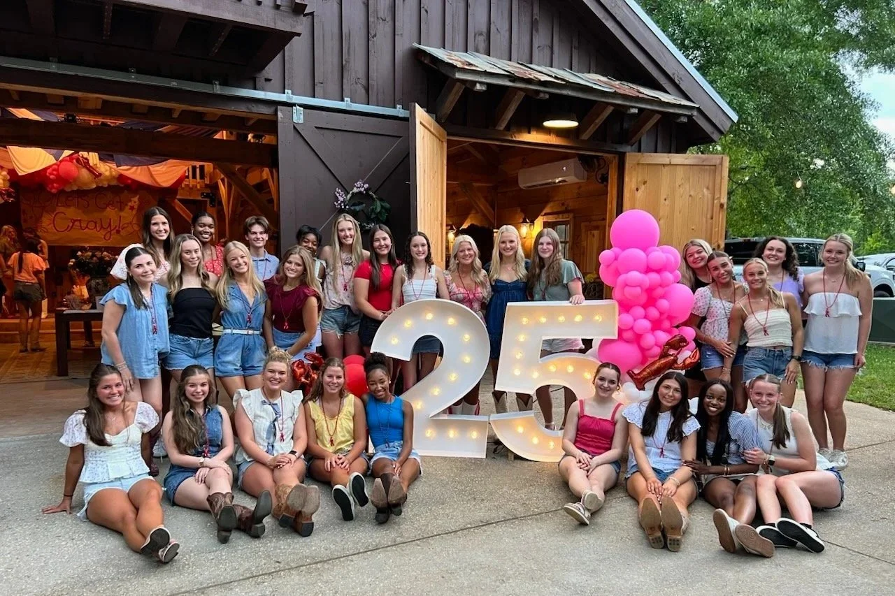 Group of young girls celebrating a 25th birthday outdoors in front of a rustic barn with large illuminated '25' sign, pink balloons, and a variety of casual summer outfits.