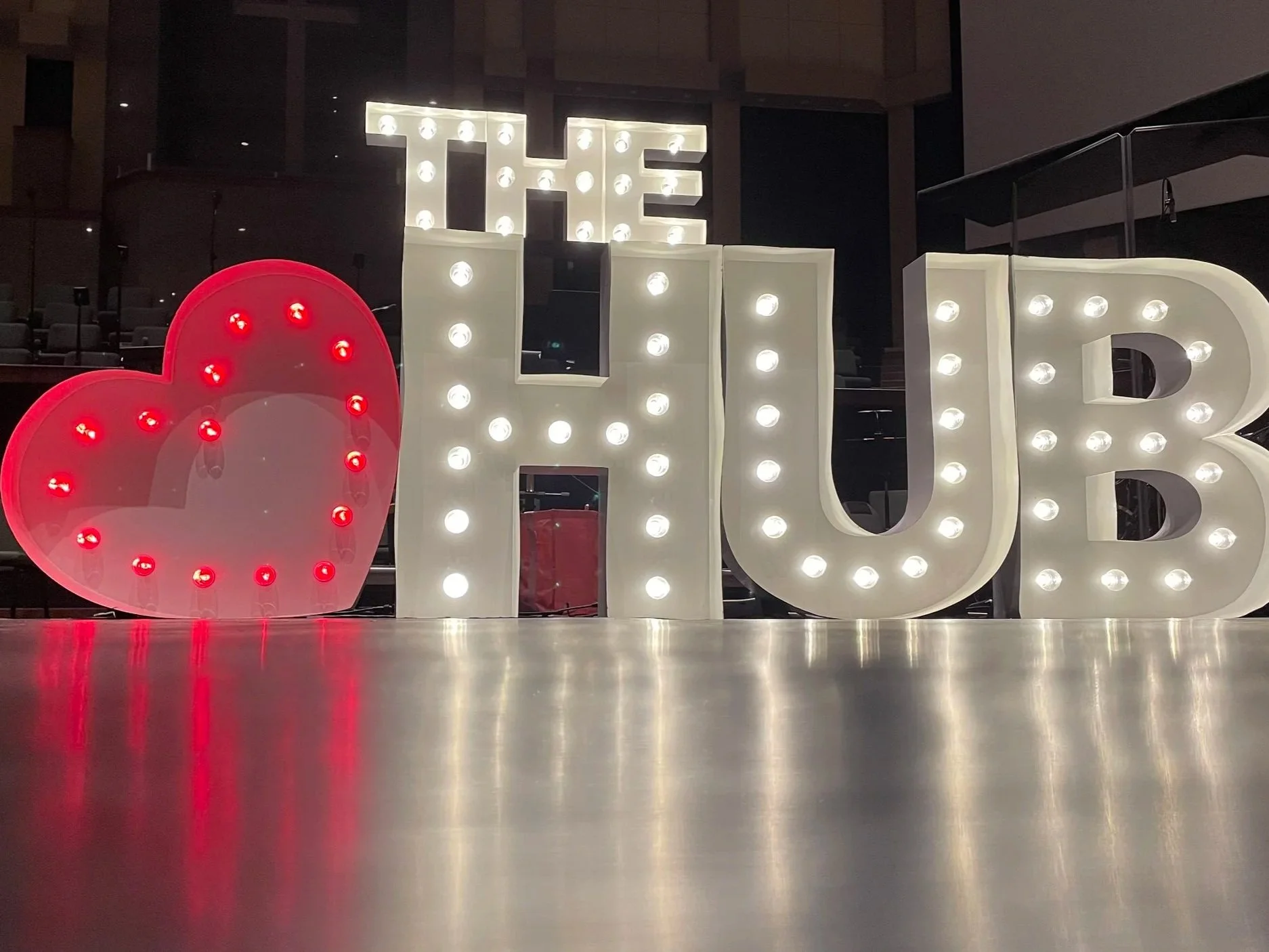 Illuminated decorative sign spells out 'Heart The HUB' with the '♥️' as the word Heart, positioned on a shiny reflective surface.