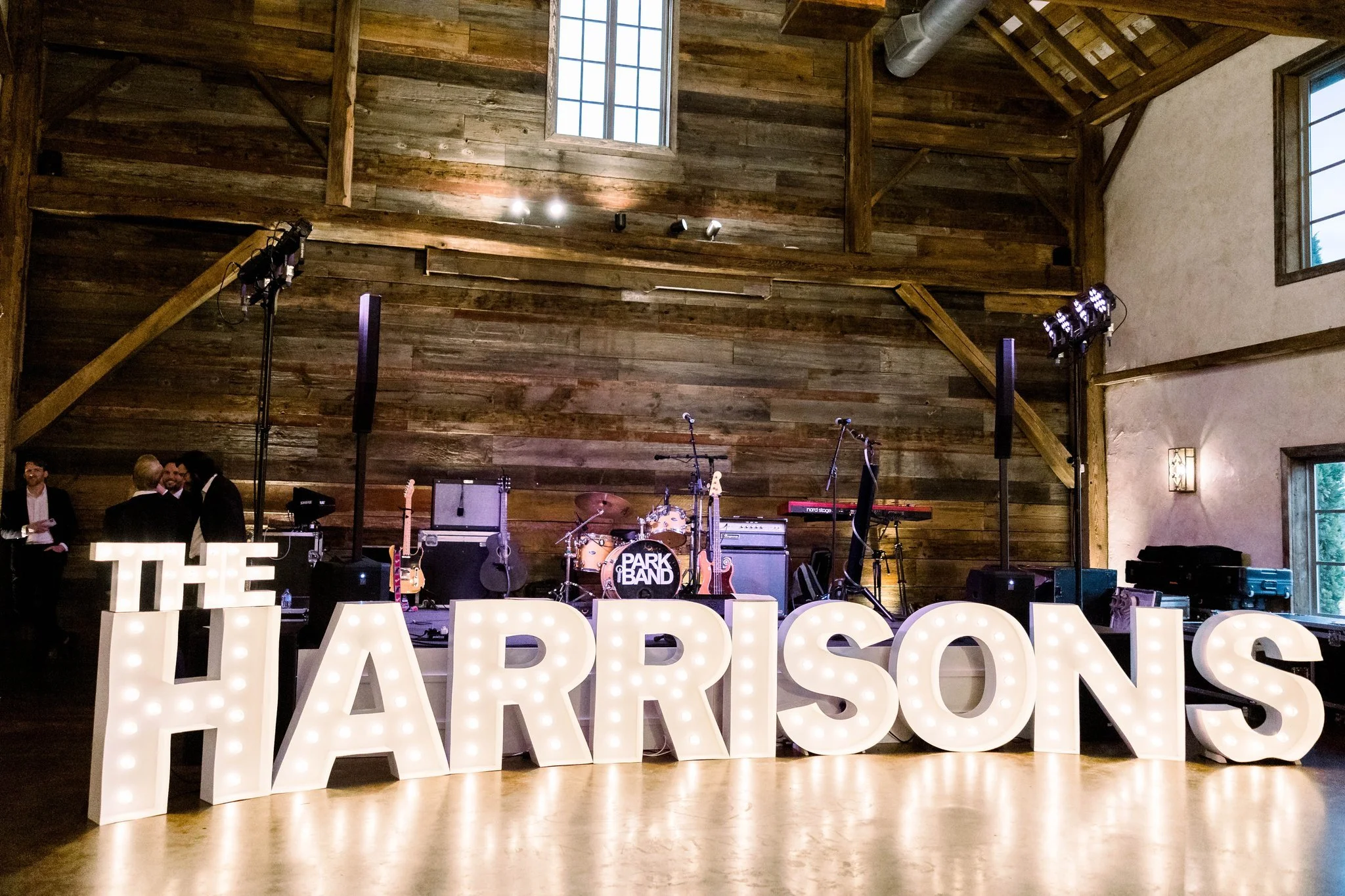 Indoor stage with musical instruments such as guitars and drums, large illuminated letters spelling 'THE' and 'HARRISONS' in front, and a rustic wood wall with windows in the background. The lighting is dim, suitable for a performance or event.