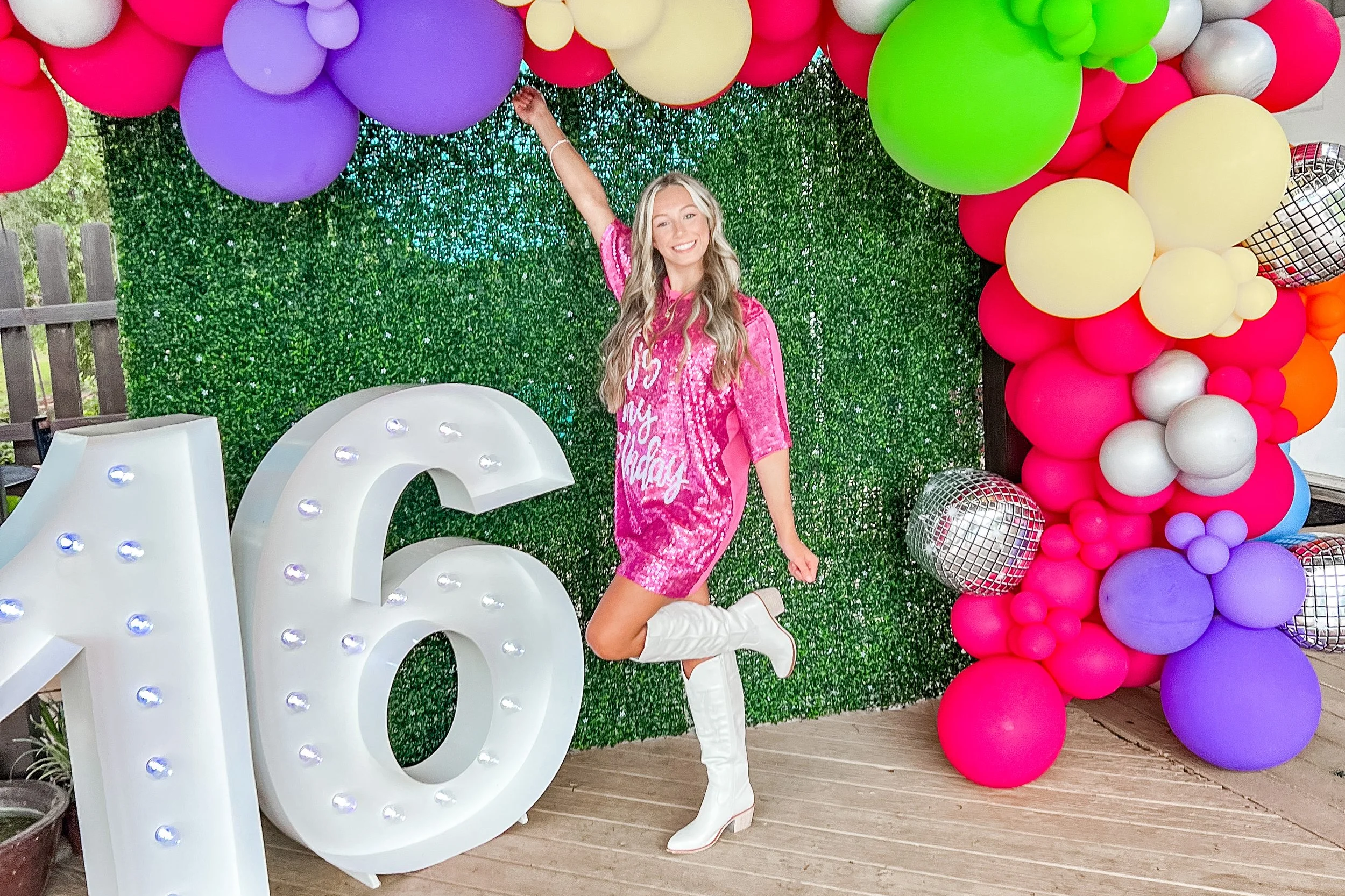 A woman in a pink sequin dress and white boots is posing and smiling in front of a large green leaf backdrop with colorful balloon decorations, including pink, purple, yellow, white, orange, and silver disco balls. Sweet 16
