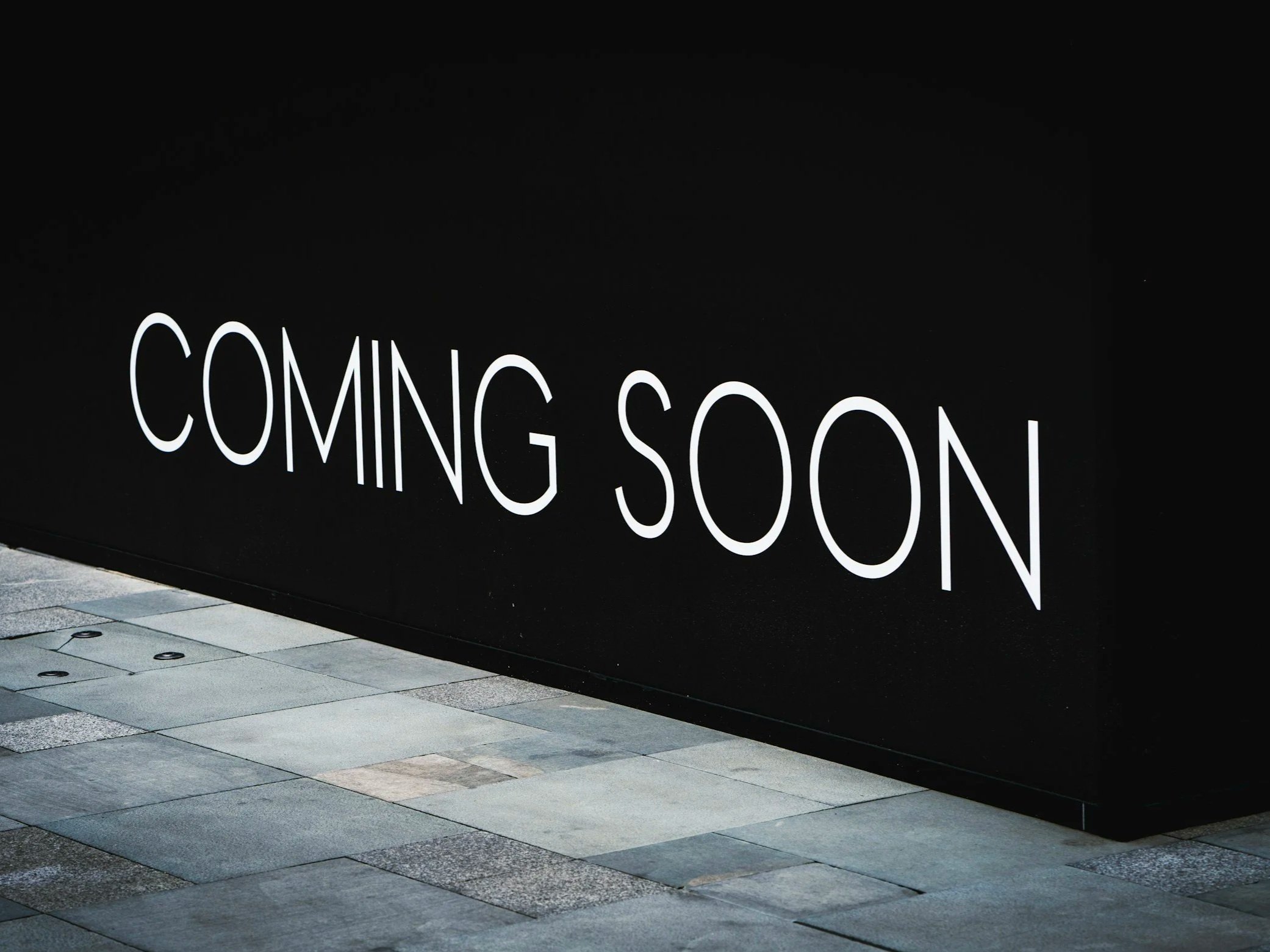 A black wall with large white text saying 'COMING SOON' and a section of tiled pavement in front.