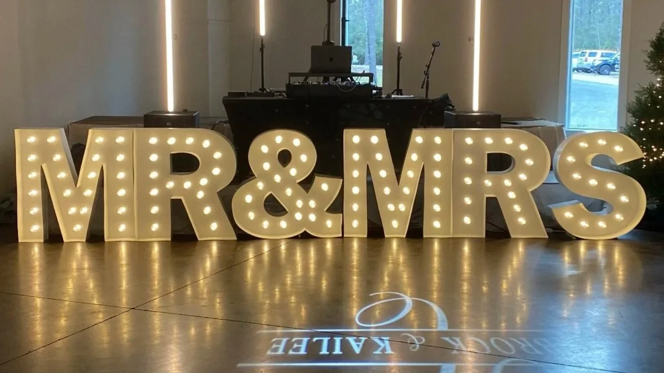 Large illuminated letters spelling "MR & MRS" on a polished floor, with a DJ setup behind and a window in the background.