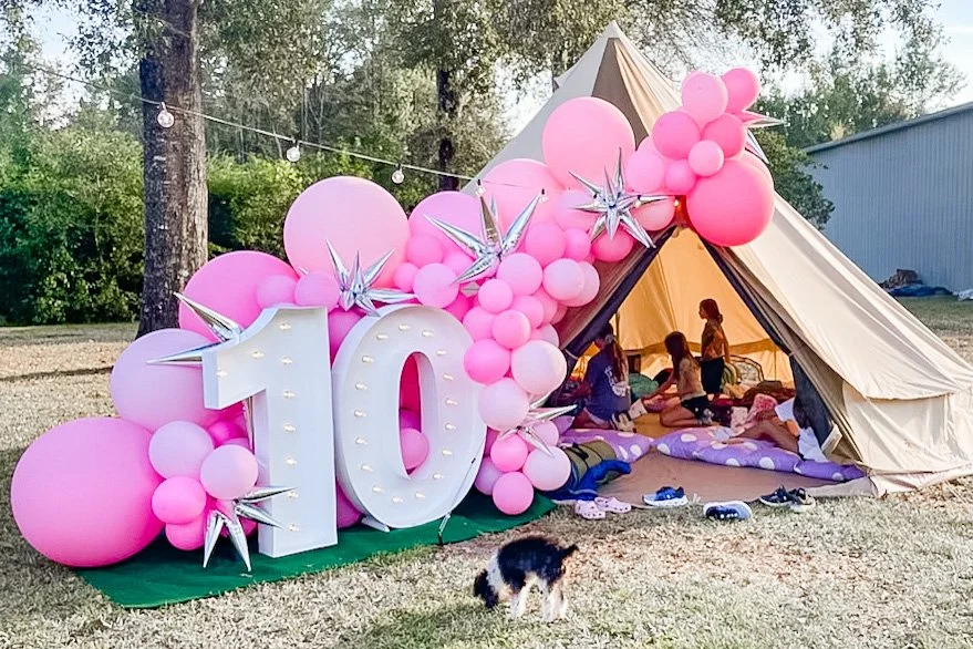 Children inside a tent decorated with pink balloons and silver star-shaped balloons, celebrating a 10th birthday outdoors, with a dog in the foreground.