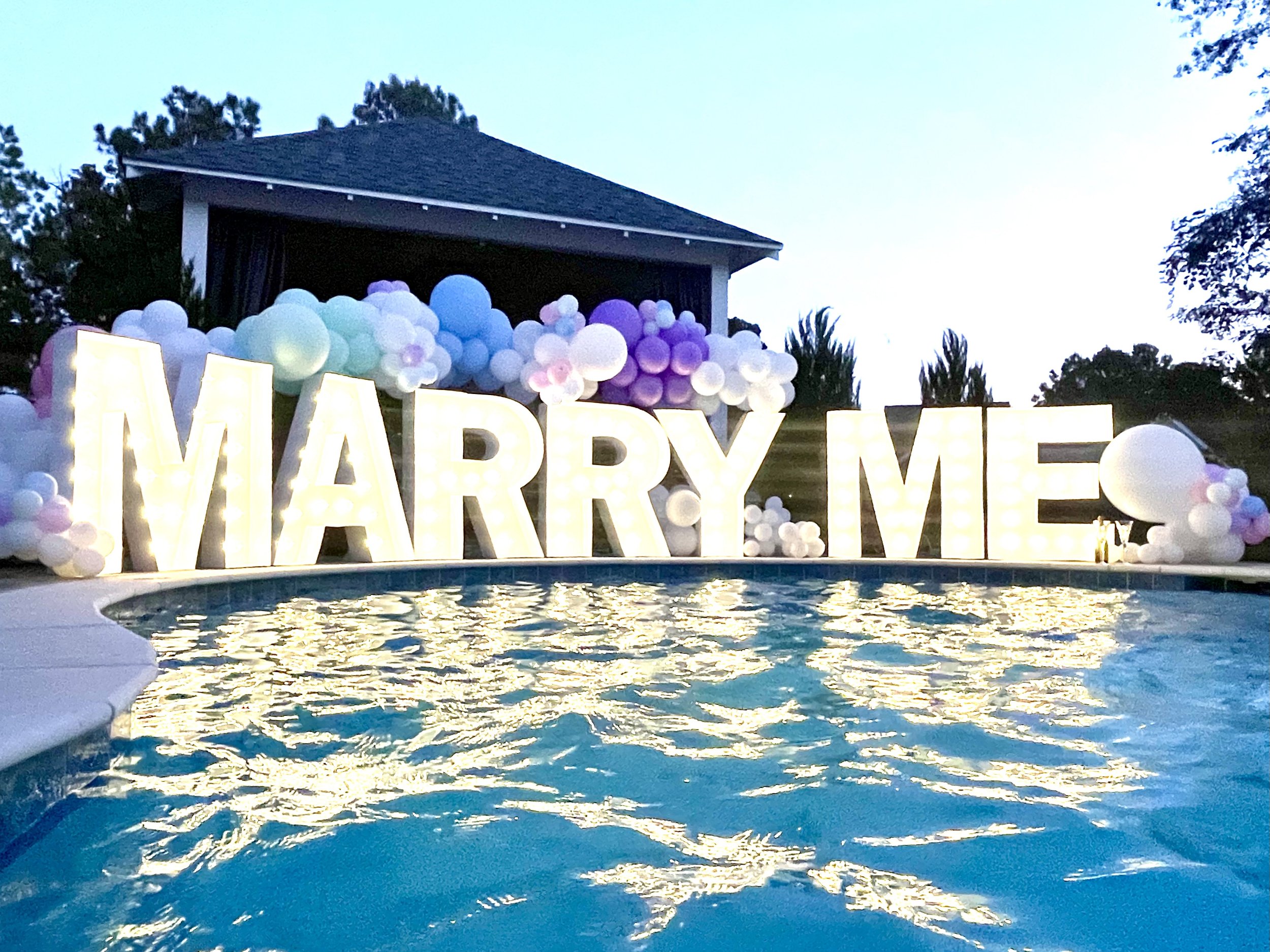 Illuminated large marquee letters spell out "MARRY ME" in front of a pool with floating candles, decorated with pastel-colored balloons and flowers under a dusky evening sky.