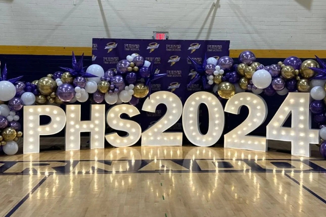 Large marquee letters spelling 'PHS 2024' with decorative balloons in purple, white, and gold in a gymnasium setting.