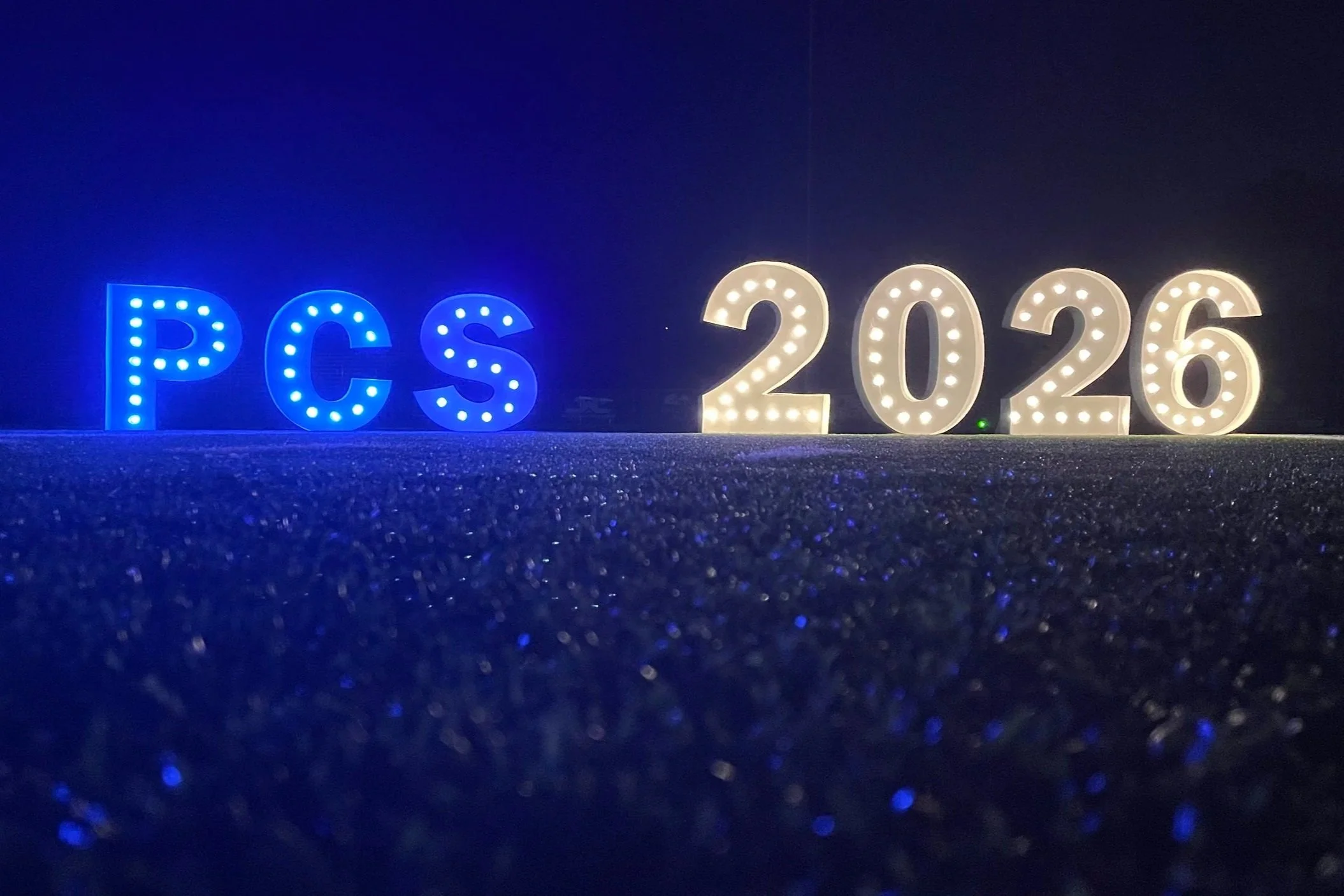 Decorative letters spelling 'PCS' illuminated in blue lights and '2026' illuminated in white lights, set against a dark background.