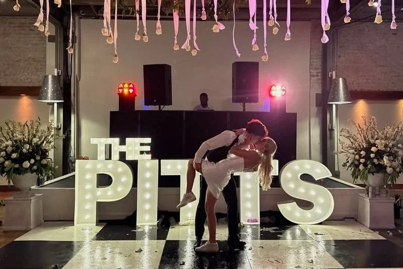 A couple dancing in front of illuminated letters spelling 'THE PITTS' at a celebration, with flowers and event lighting in the background.